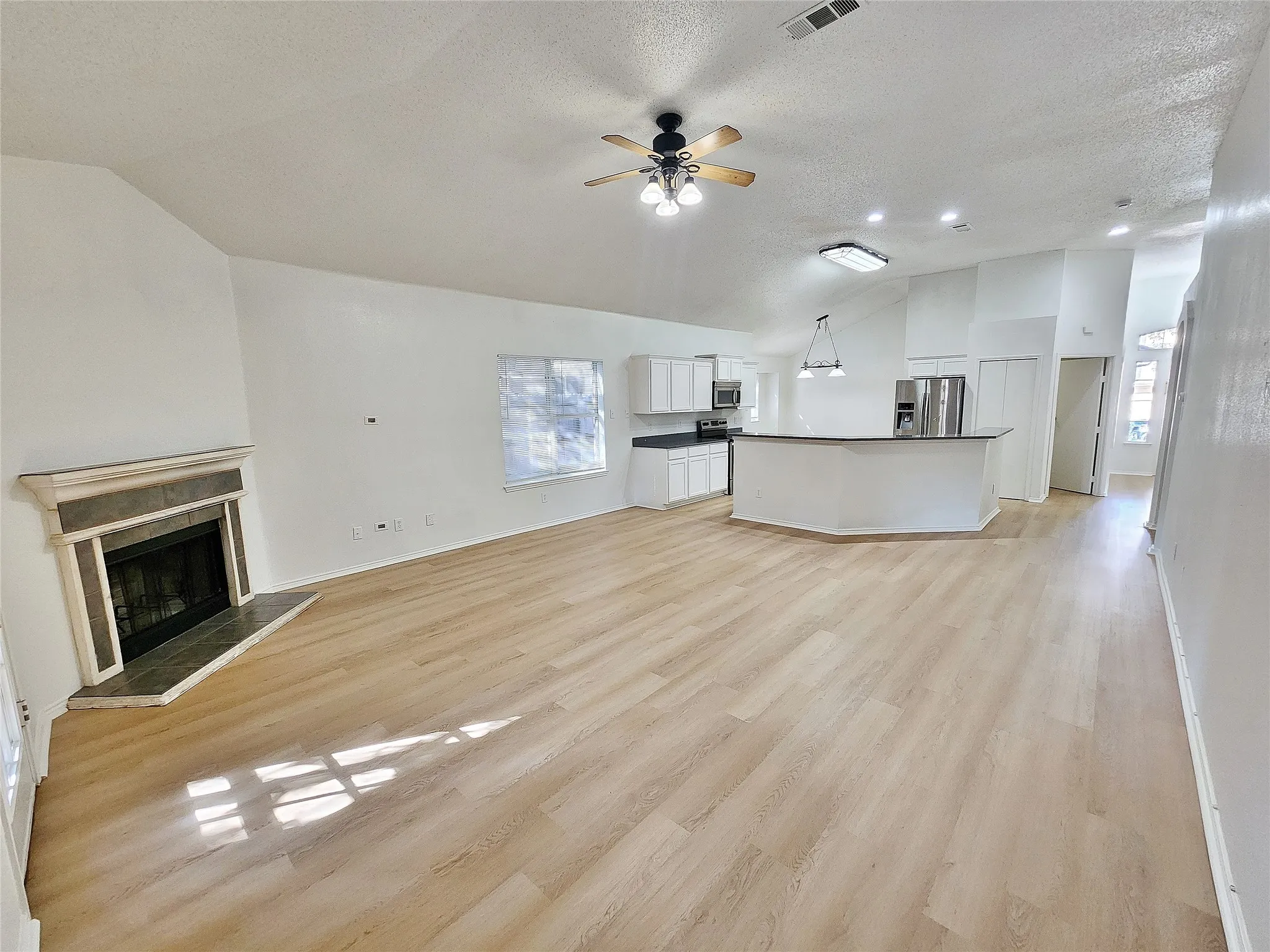 Open-concept living area featuring wood-finish flooring, a fireplace with a stone surround, and an integrated kitchen with white cabinetry and a breakfast bar