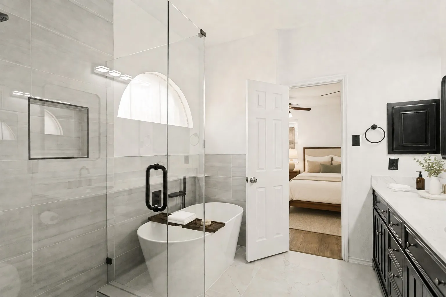 Freestanding soaking tub with a wall-mounted faucet, glass-enclosed shower featuring a tiled niche, double vanity with a light-toned countertop, and dark wood-finish cabinetry