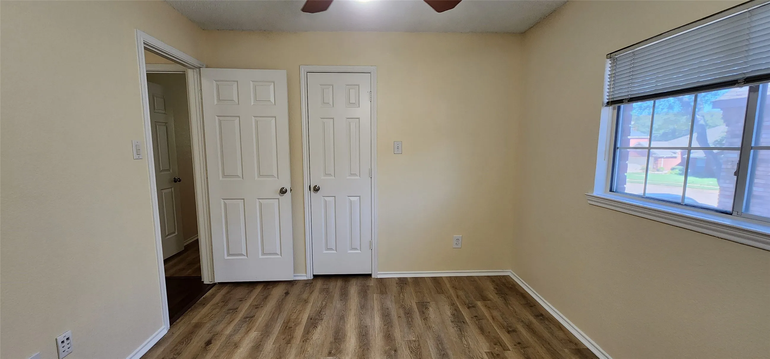 Wood-finish flooring, light-toned wall paint, ceiling fan, white paneled doors, and a window with blinds
