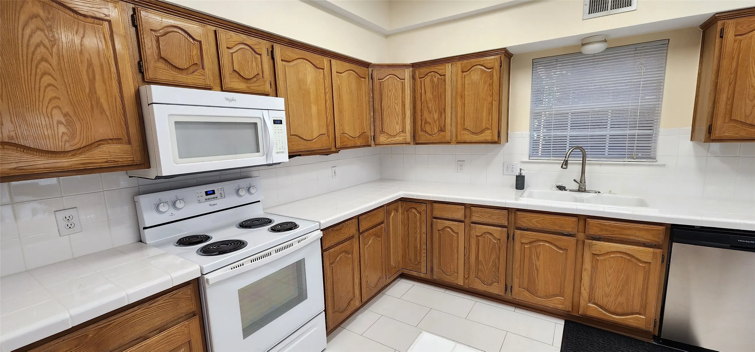 Wood-finish cabinetry, white tile countertops, white tile backsplash, stainless steel sink, and stainless steel dishwasher