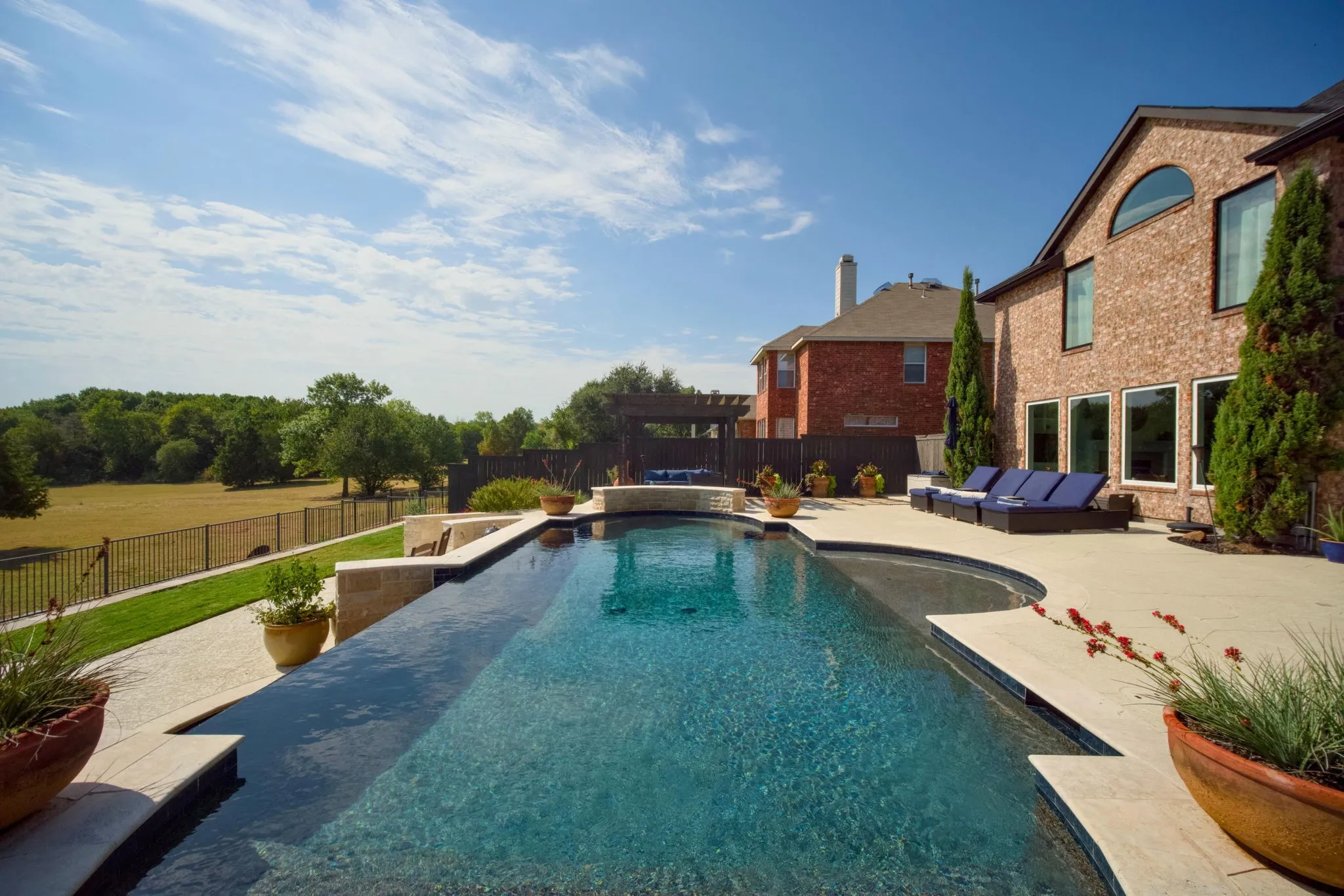 Infinity-edge swimming pool with an integrated spa, expansive paver patio, brick exterior, and a manicured lawn