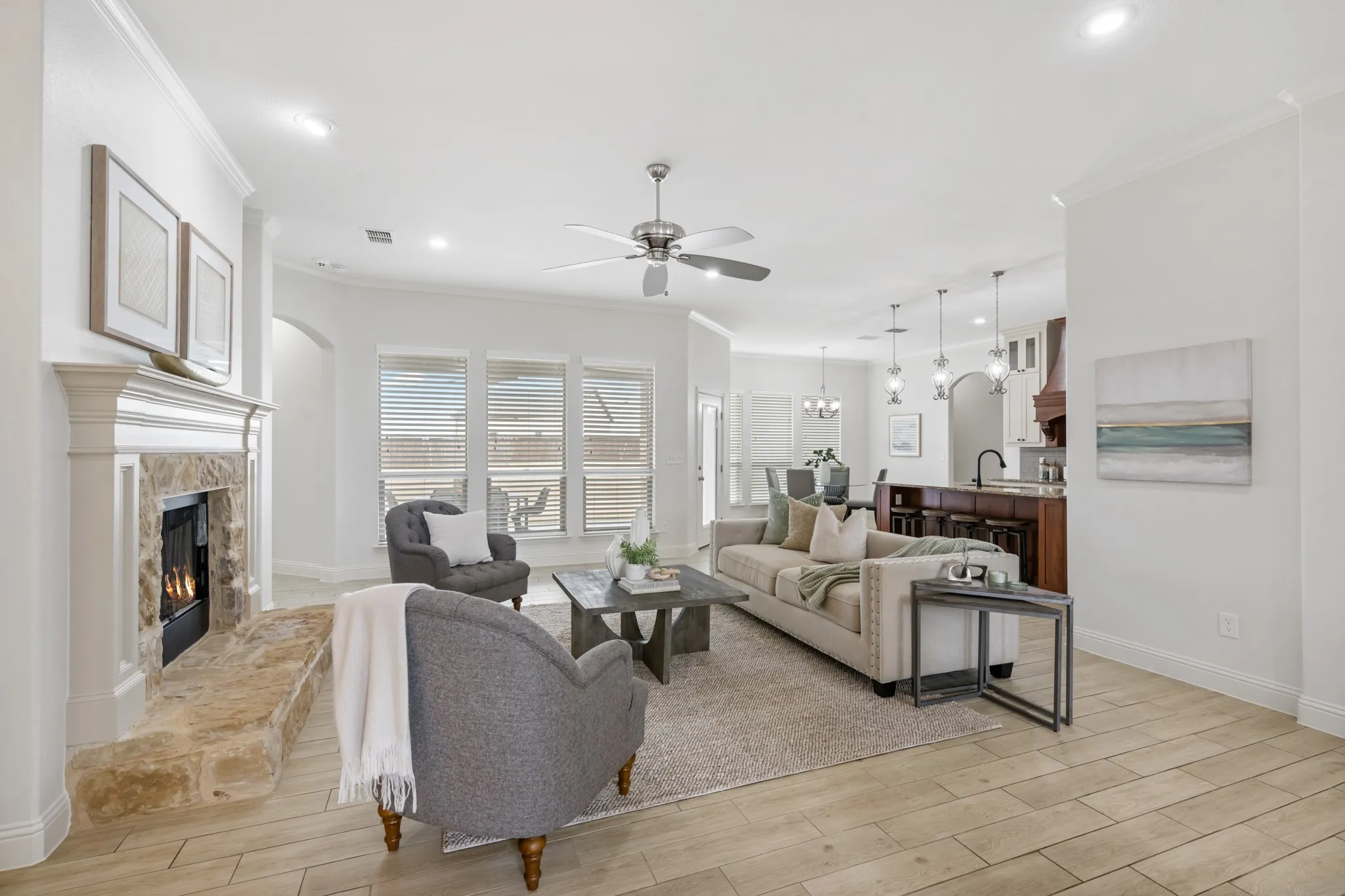 Bright and spacious open-concept living area featuring wood-finish flooring throughout, a gas fireplace with surround millwork, recessed and pendant lighting, a ceiling fan, and large multi-pane windows with plantation shutters