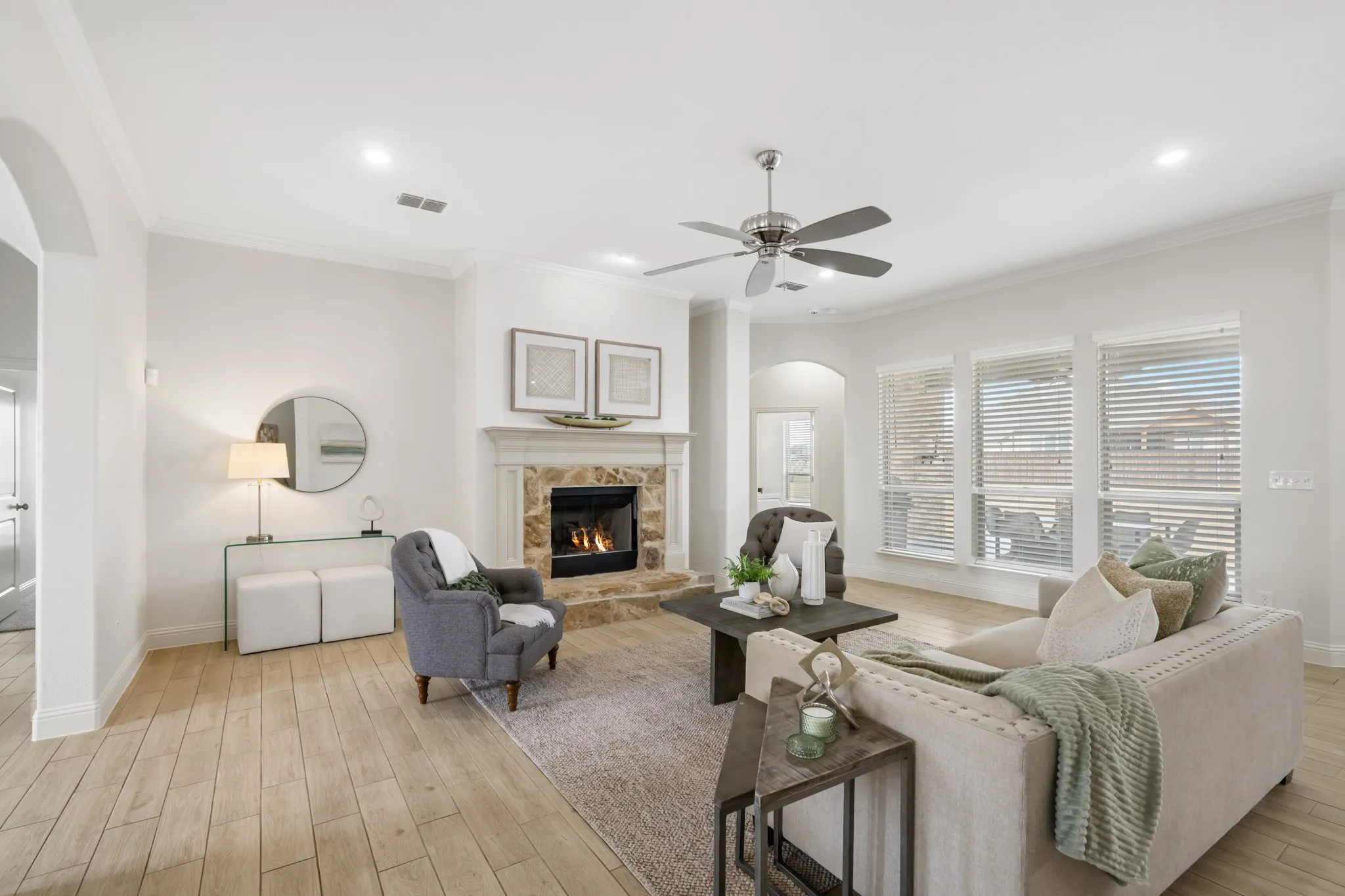 Living area featuring wood-finish flooring, a stone-surround fireplace with mantel, ceiling fan, crown molding, and multiple windows with blinds