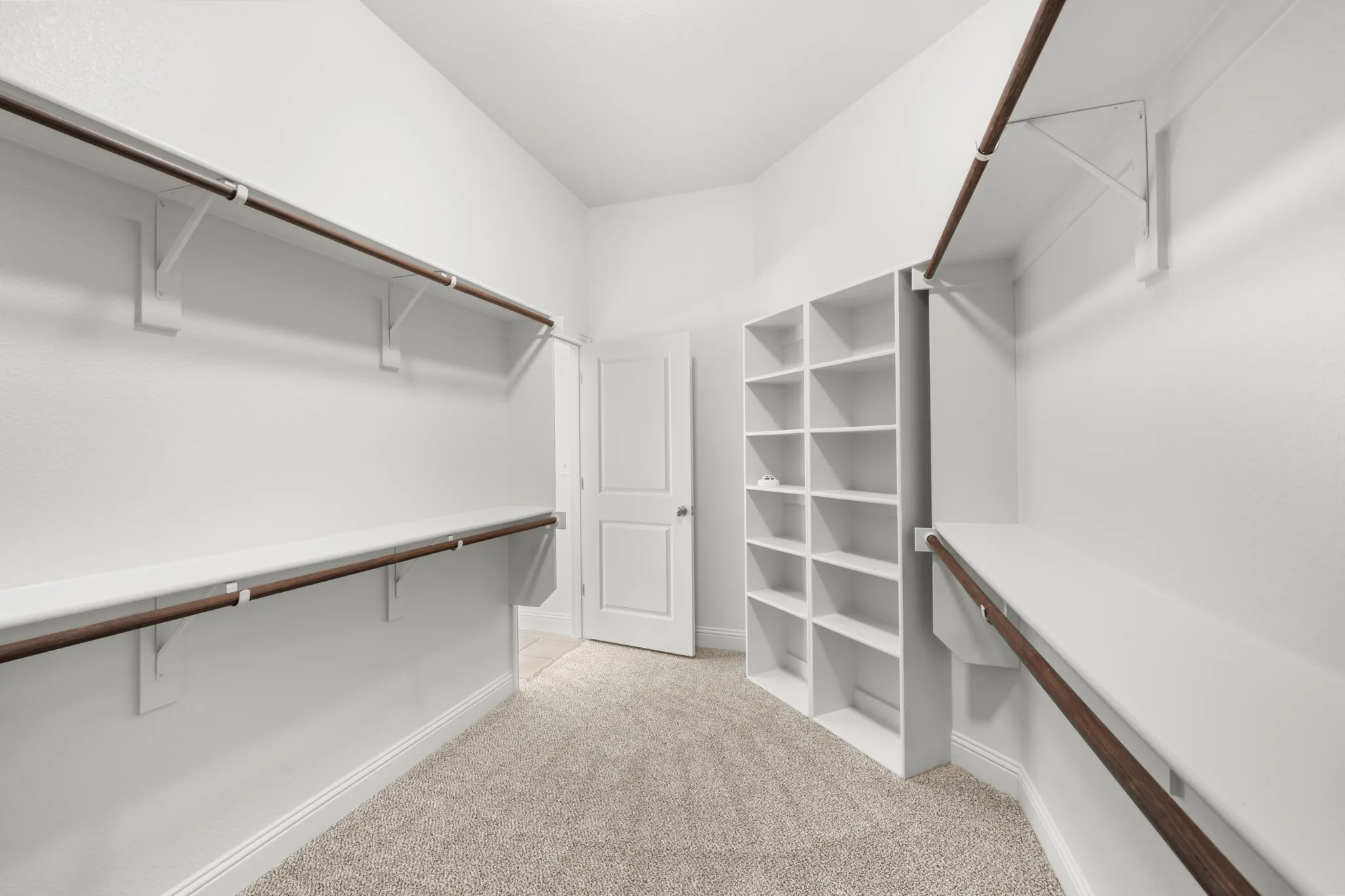 Spacious walk-in closet featuring dual clothing rods with metal finishes, white built-in shelving with multiple compartments, soft neutral carpet flooring, and abundant natural lighting from recessed fixtures