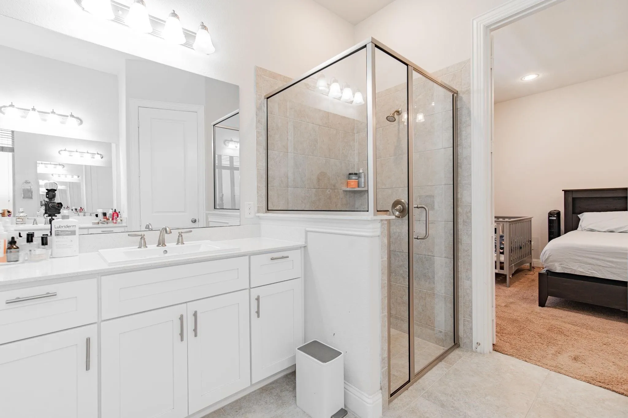 White vanity with dual sinks and brushed nickel fixtures, glass-enclosed shower with tiled surround, and ceramic tile flooring