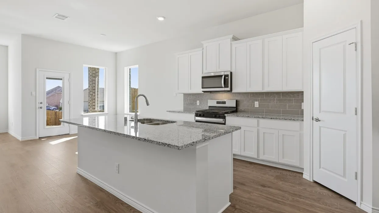 Modern kitchen featuring white shaker-style cabinetry, a large waterfall-edge island with granite countertop, stainless steel appliances including a microwave and cooktop, subway tile backsplash, and wood-finish flooring that extends into the adjoining living area with abundant natural light from multiple windows and glass doors