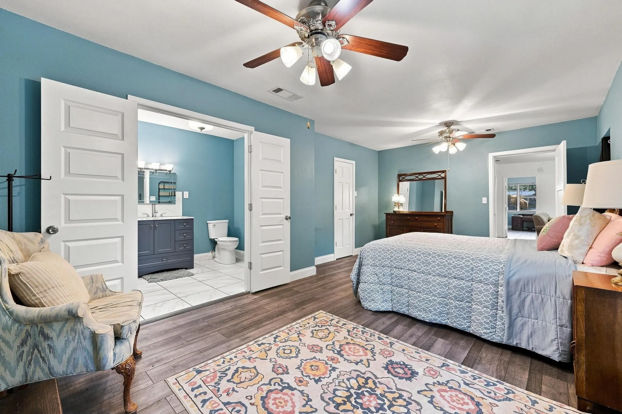 Spacious bedroom with wood-finish flooring, two ceiling fans, and an attached bathroom featuring a single vanity and tile flooring