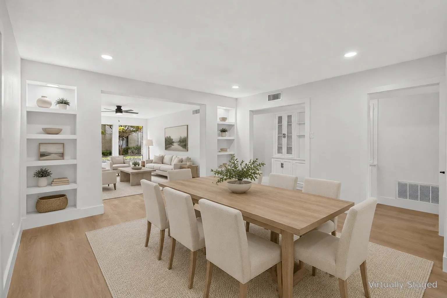 Open-concept dining and living area featuring a wood-finish dining table, white cabinetry with glass-front doors, recessed ceiling lighting, and hardwood flooring throughout