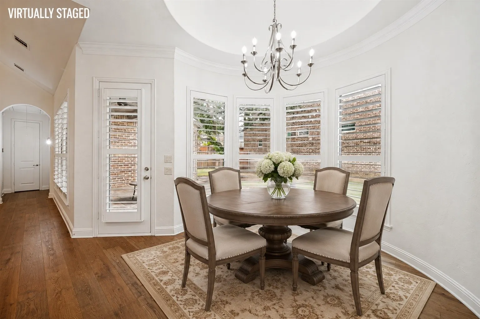 Enjoy your morning coffee in this light-filled dining space, featuring a dramatic tray ceiling, elegant chandelier, plantation shutters, and a wall of windows that fill the space with natural light.