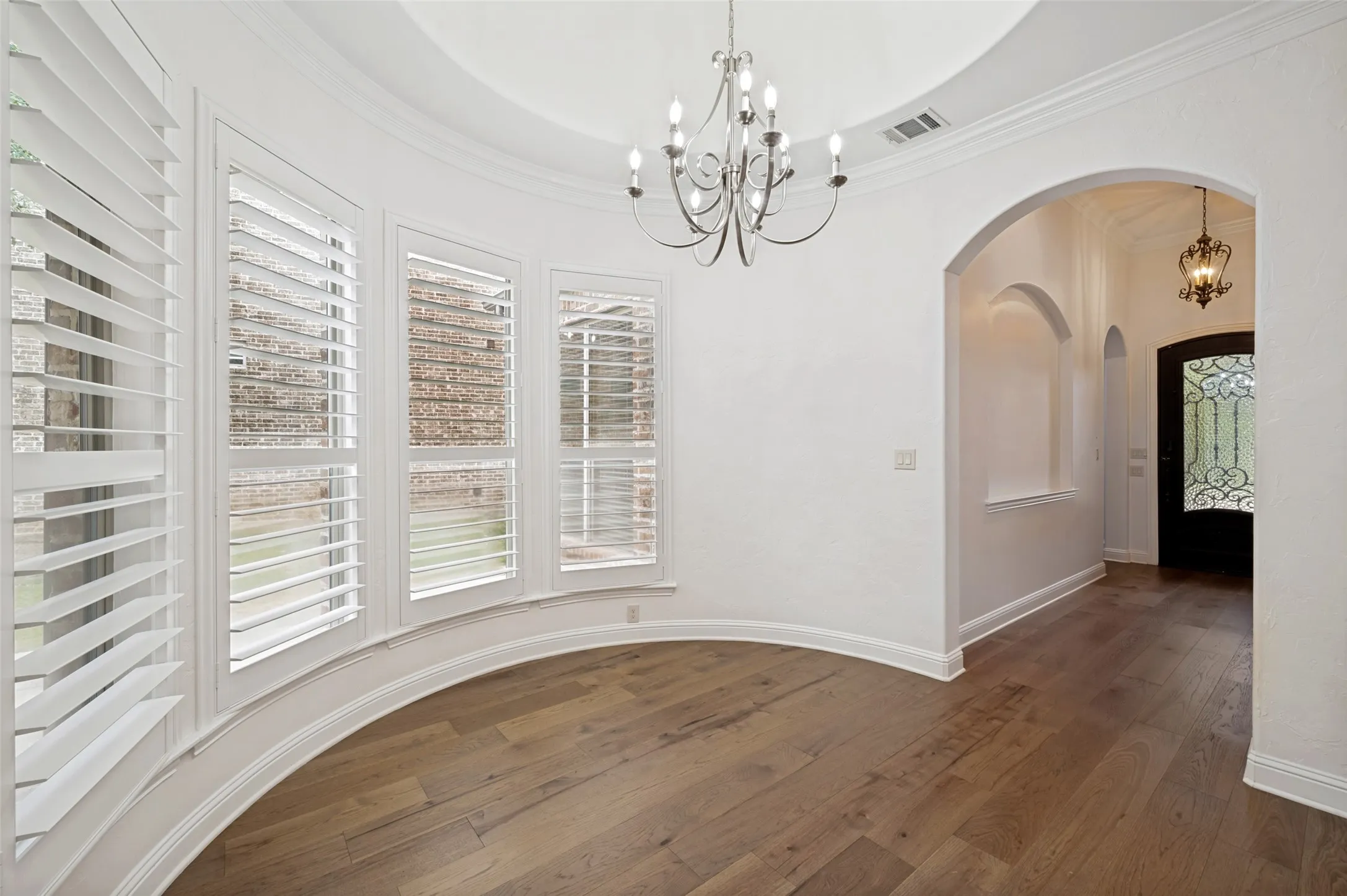 The elegant dining space welcomes you with a dramatic tray ceiling, designer chandelier, plantation shutters, and graceful curved windows, creating the perfect space for casual dining while offering a beautiful view of the grand foyer.