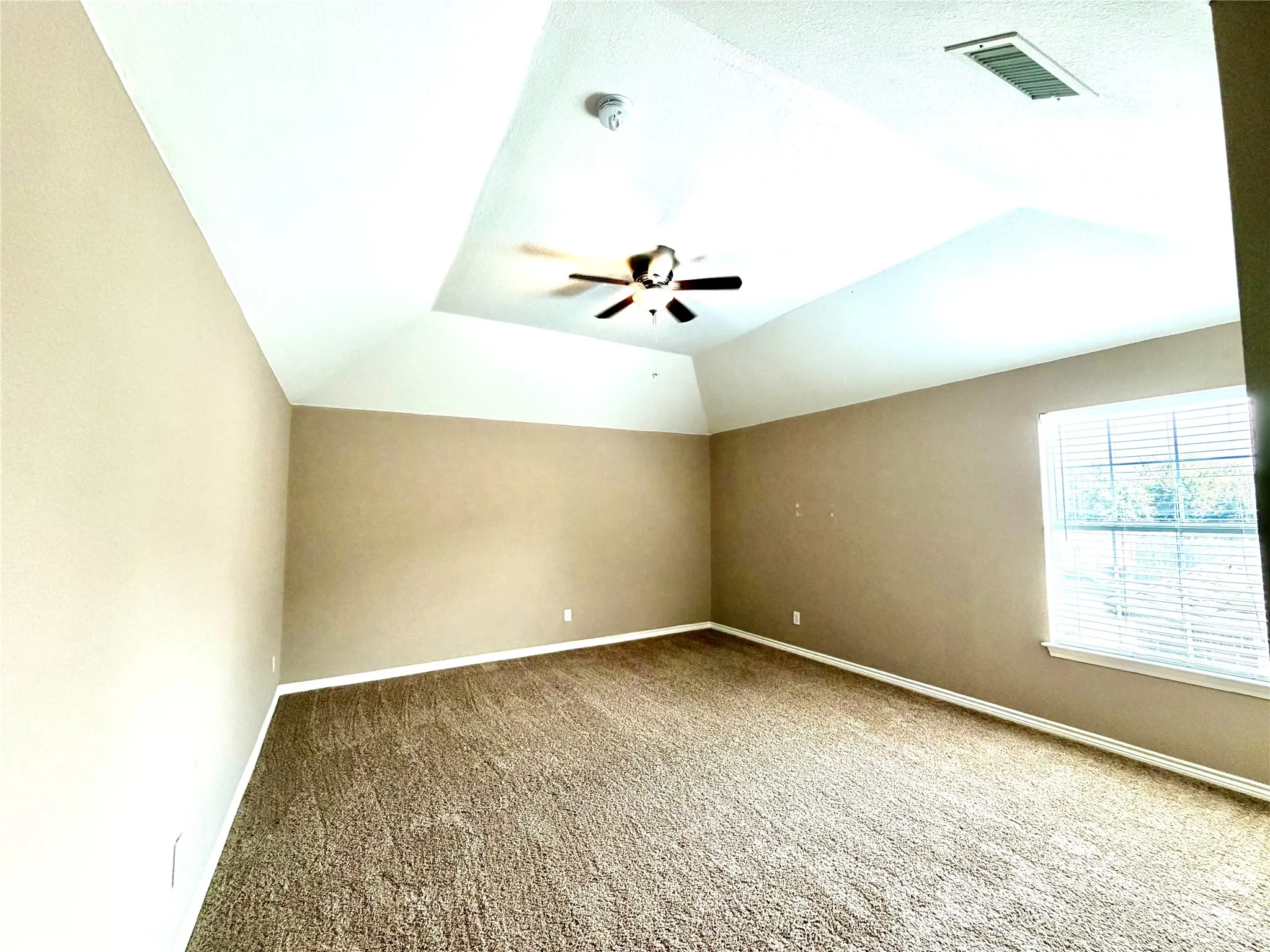 Vaulted tray ceiling with a built-in ceiling fan, neutral carpet flooring, and a window with blinds
