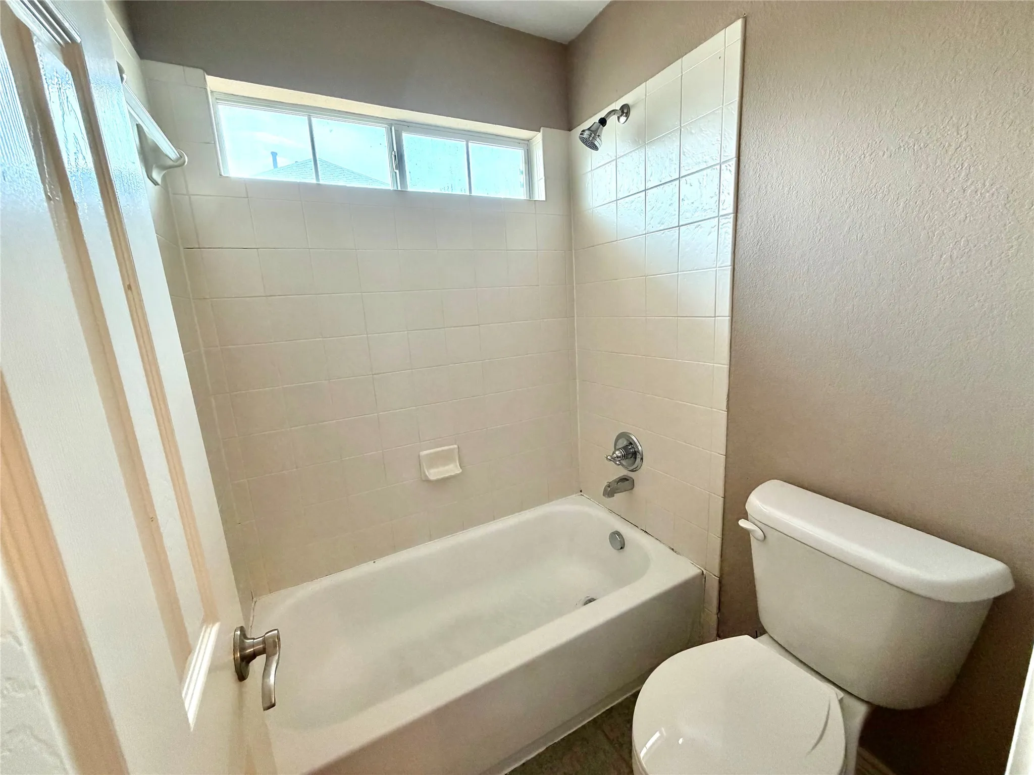 White porcelain bathtub with tile surround, chrome fixtures, and an upper transom window
