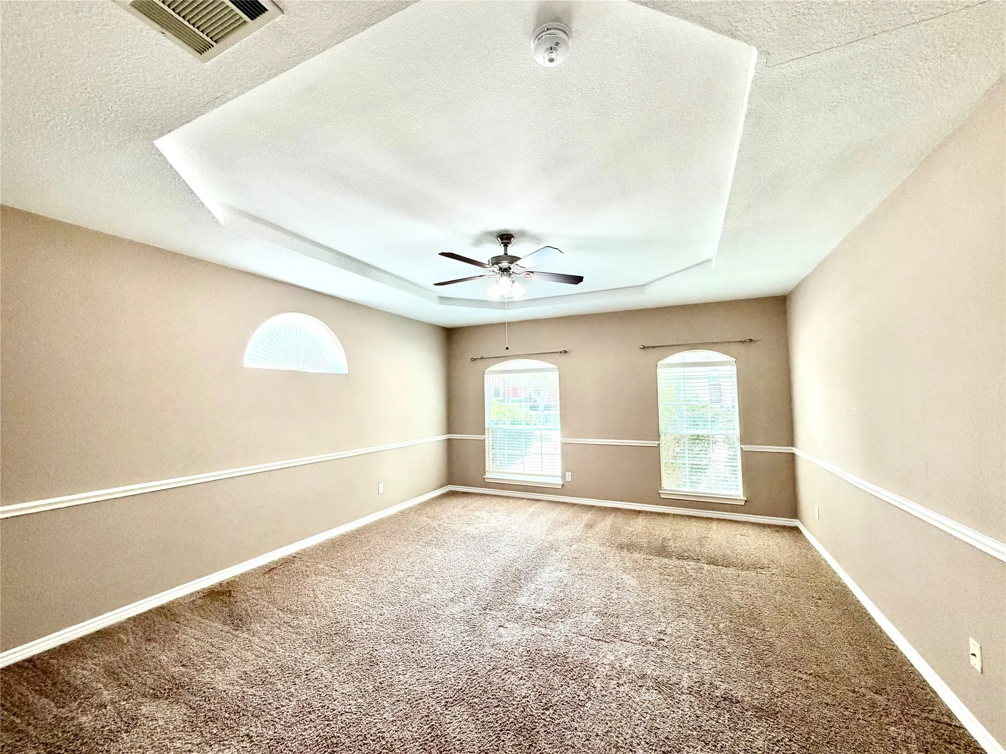 Tray ceiling with a ceiling fan, arched transom window, two arched windows, and wall trim