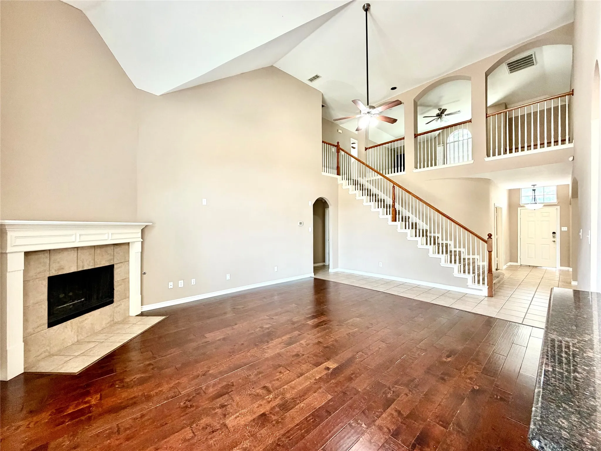 Vaulted ceiling with a ceiling fan, fireplace with a tile surround and mantel, wood-finish flooring, staircase with wood handrails, and a tiled entryway