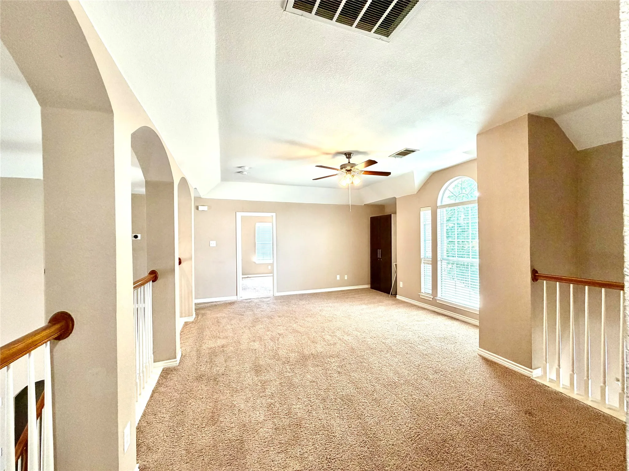 Spacious carpeted room featuring a ceiling fan, arched windows with blinds, and white balusters with a wood handrail