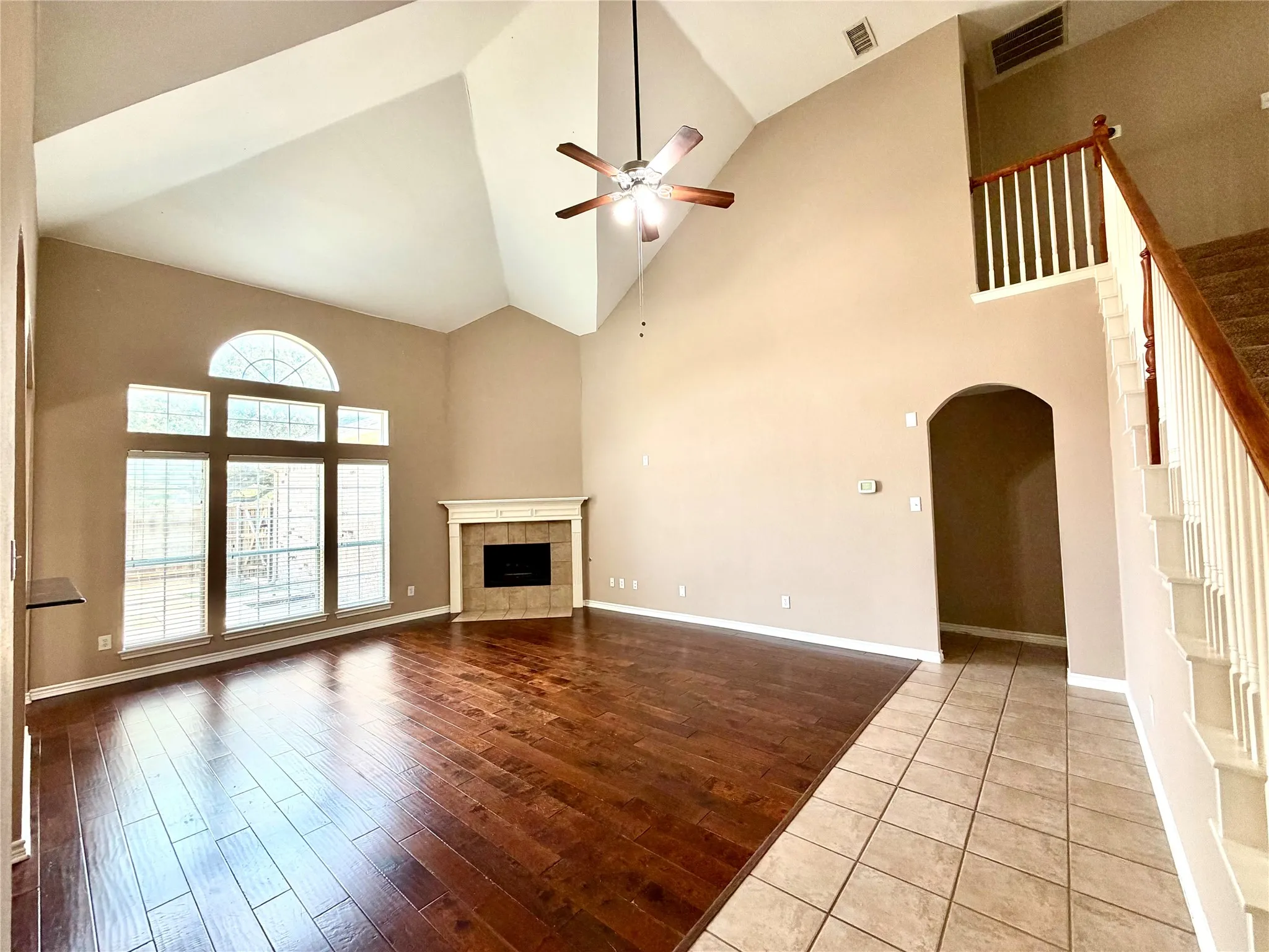 Soaring vaulted ceilings with a ceiling fan, expansive windows including an arched transom, a tiled fireplace with a mantel, dark wood-finish flooring, and a tiled entryway