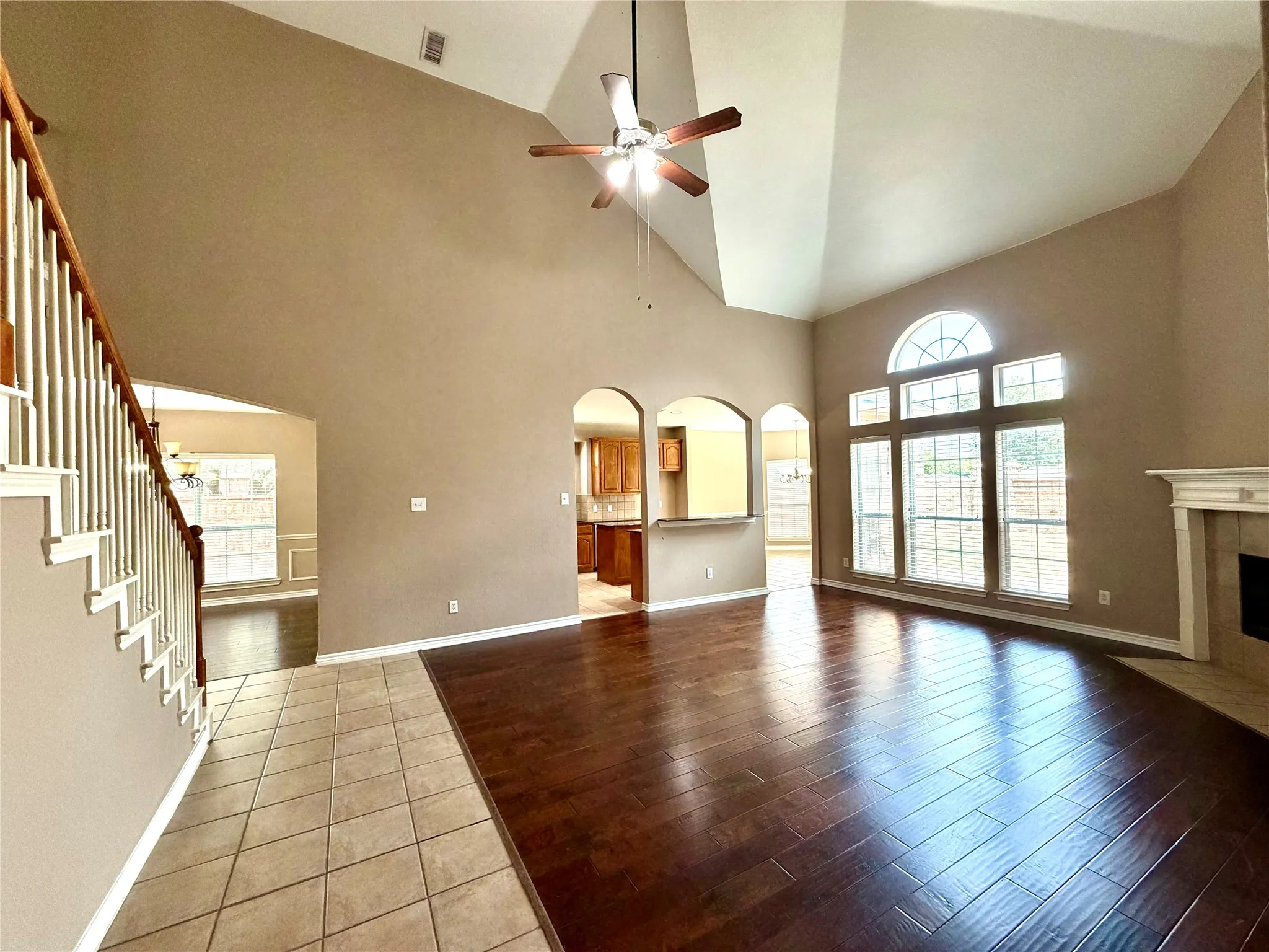 Vaulted ceiling with a ceiling fan, arched windows with an arched transom, a white mantel, wood-finish flooring, and a staircase with a wood handrail