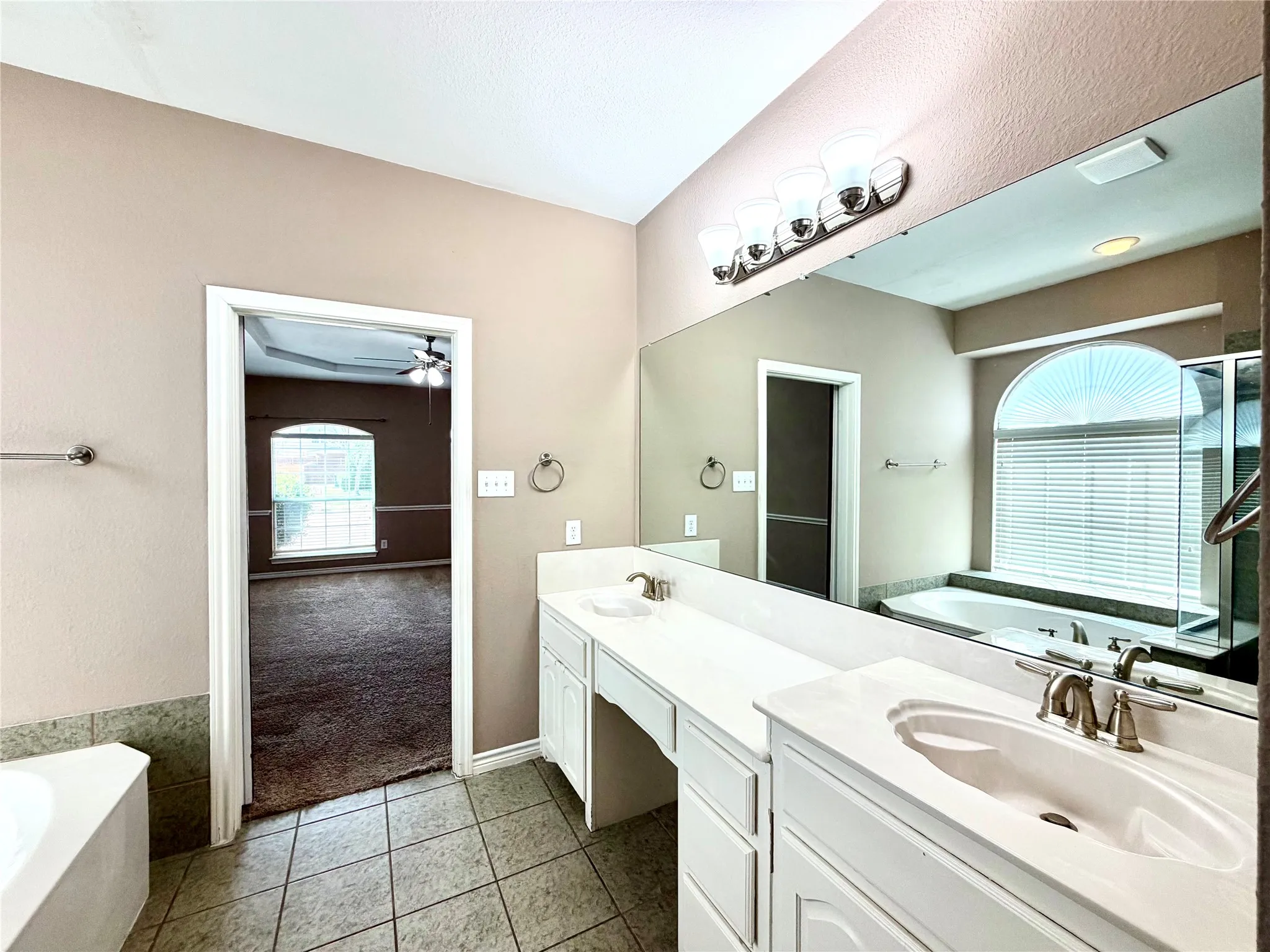 Dual vanity with integrated sinks and white cabinetry, expansive wall-mounted mirror, tile flooring, built-in corner tub, and arched window with horizontal blinds