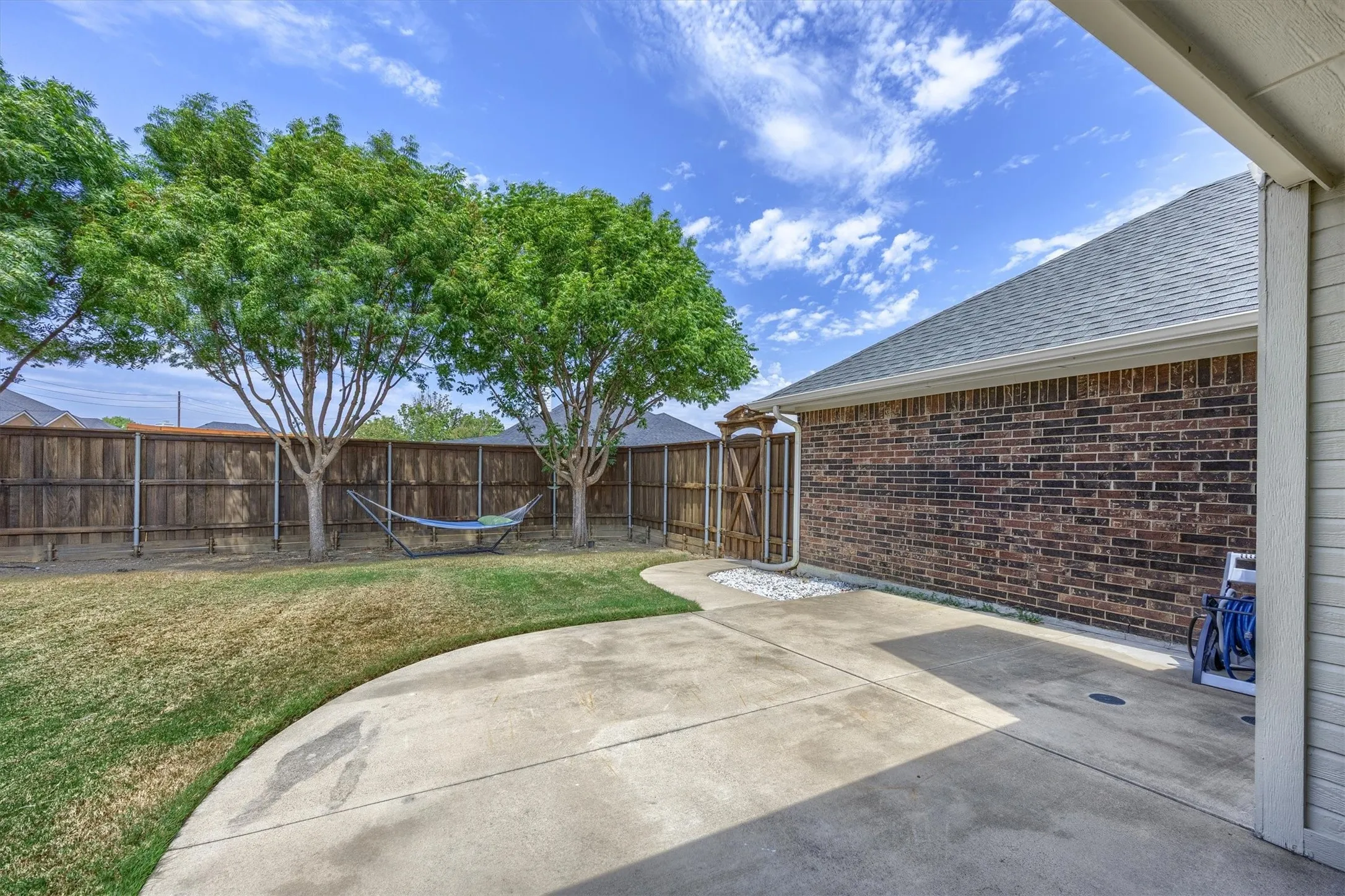 Expansive concrete patio with a brick exterior, mature trees, wood fence, and a gate