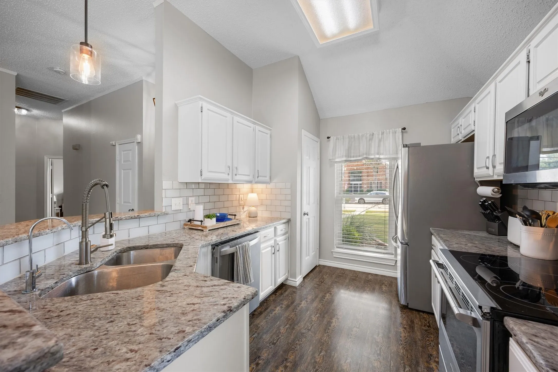 Modern kitchen featuring white cabinetry, subway tile backsplash, dual stainless steel sinks with brushed nickel faucet, granite countertops, stainless steel appliances, wood-finish flooring, and abundant natural light.