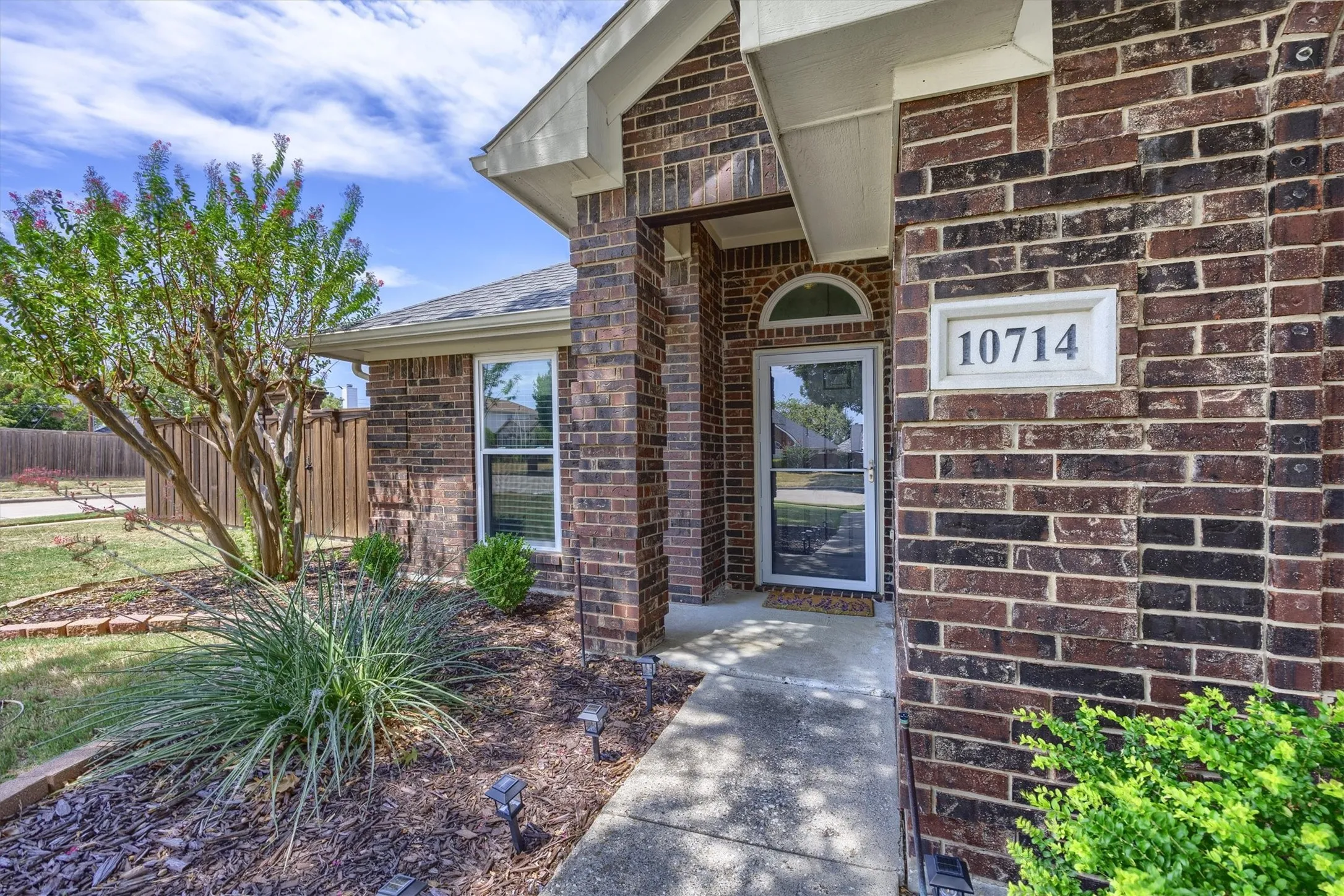 Brick exterior with white trim, arched transom above the entry door, concrete walkway, established landscaping, and a fenced perimeter