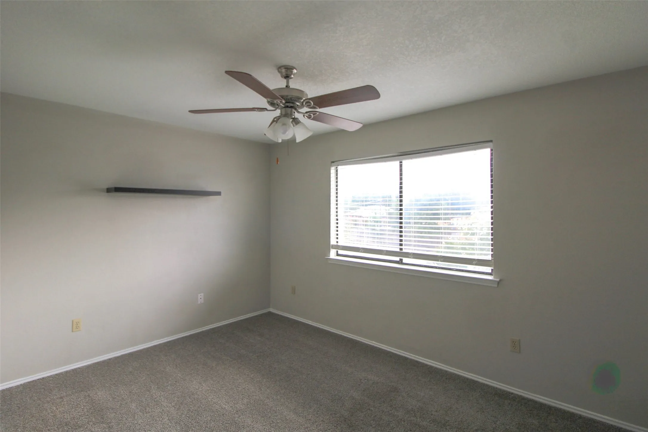 Neutral wall color, wall-to-wall carpeting, window with blinds, ceiling fan with light fixture, and floating wall shelf