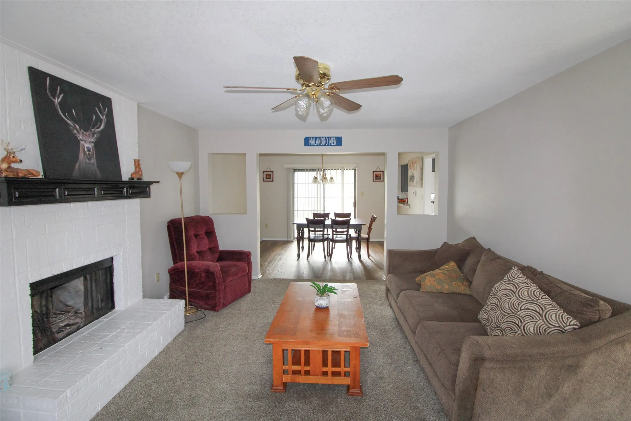 Living area featuring a brick fireplace with a mantel, carpet flooring, a ceiling fan, and an opening to a dining area with wood-finish flooring