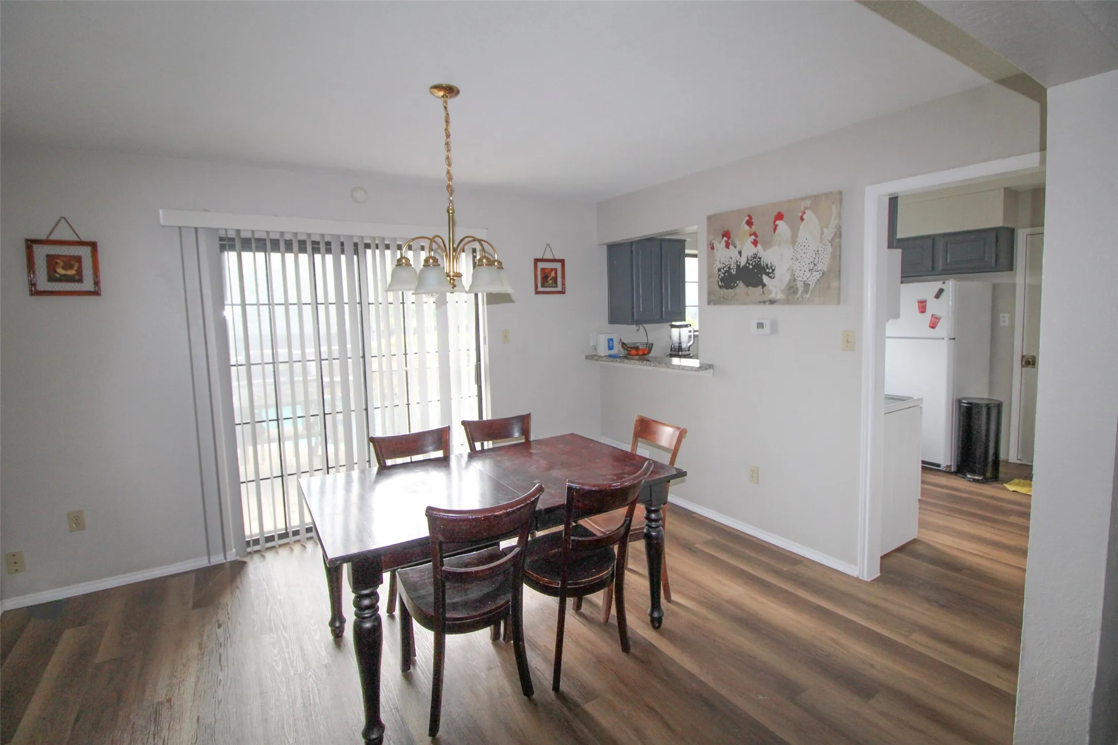 Wood-finish flooring throughout, sliding glass door with vertical blinds, brass-finish chandelier, and a pass-through to the kitchen with dark cabinetry