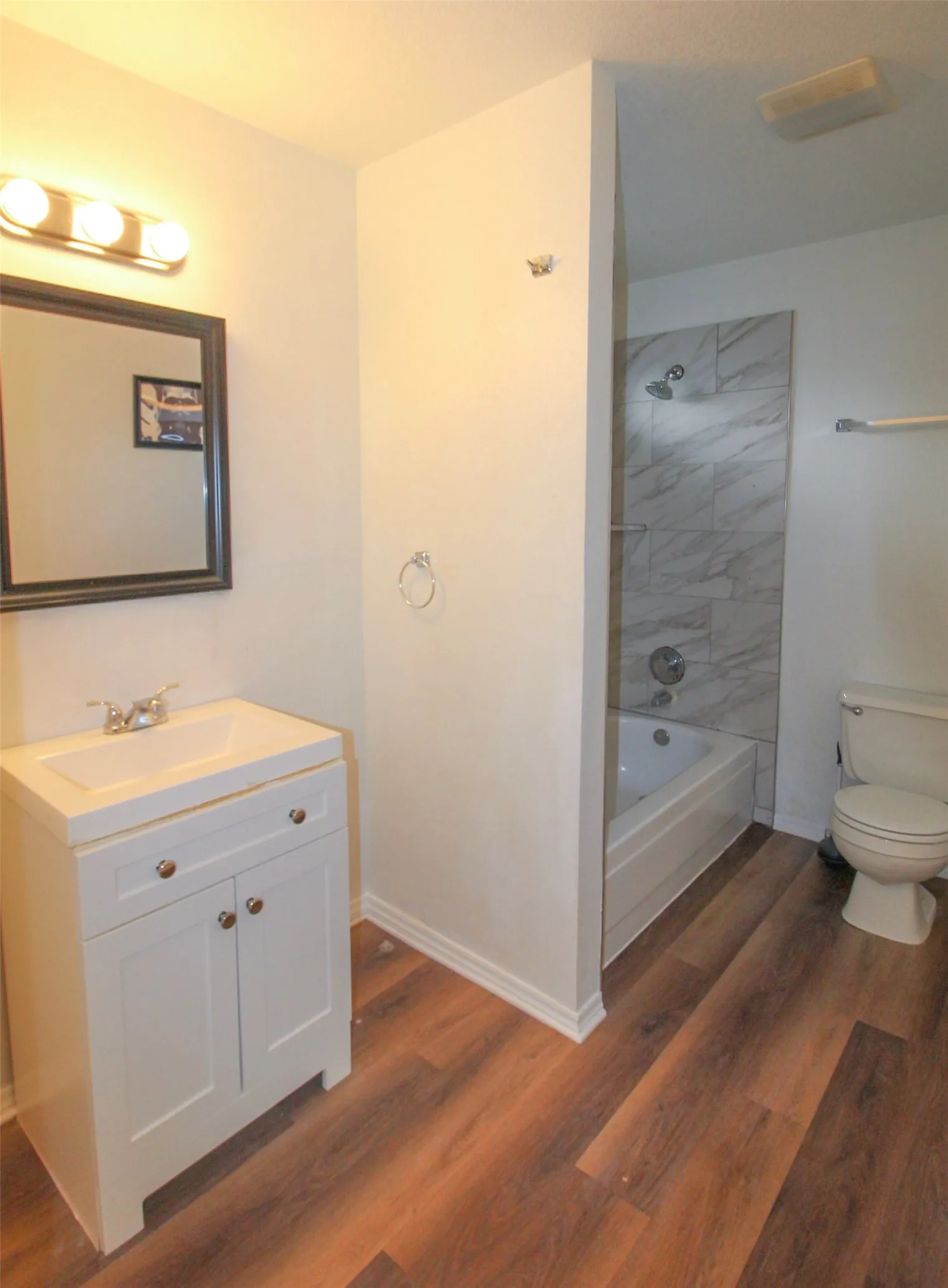 Bathroom featuring a white vanity with integrated sink, wood-finish flooring, a full bathtub with tile surround, and a toilet