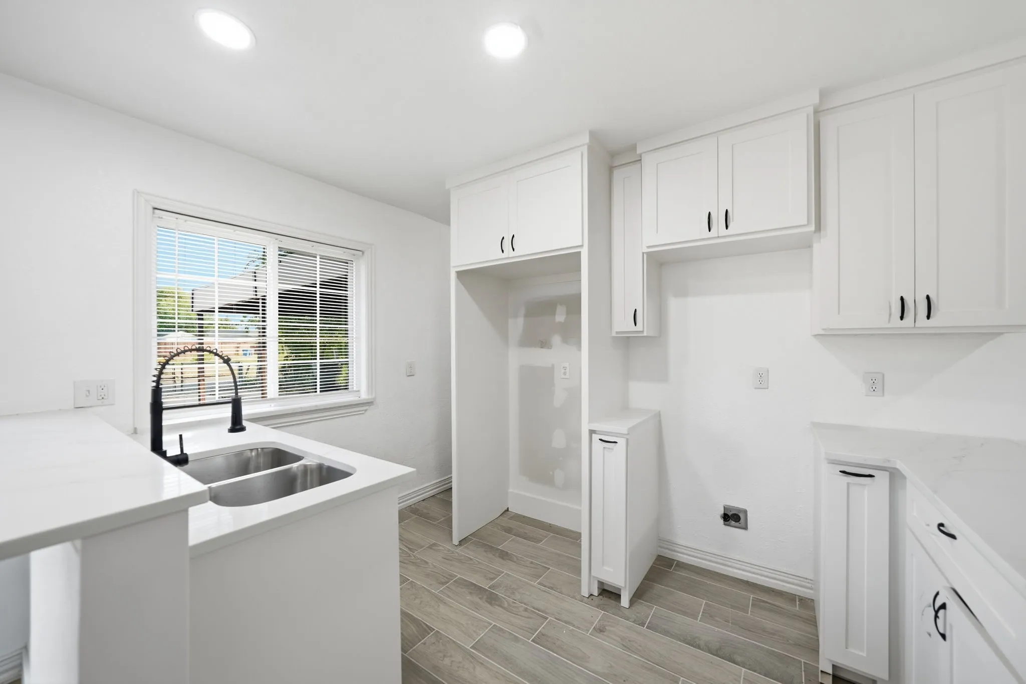 White shaker-style cabinetry with black hardware, white countertops, dual basin stainless steel sink with a black gooseneck faucet, window with blinds, and wood-finish flooring