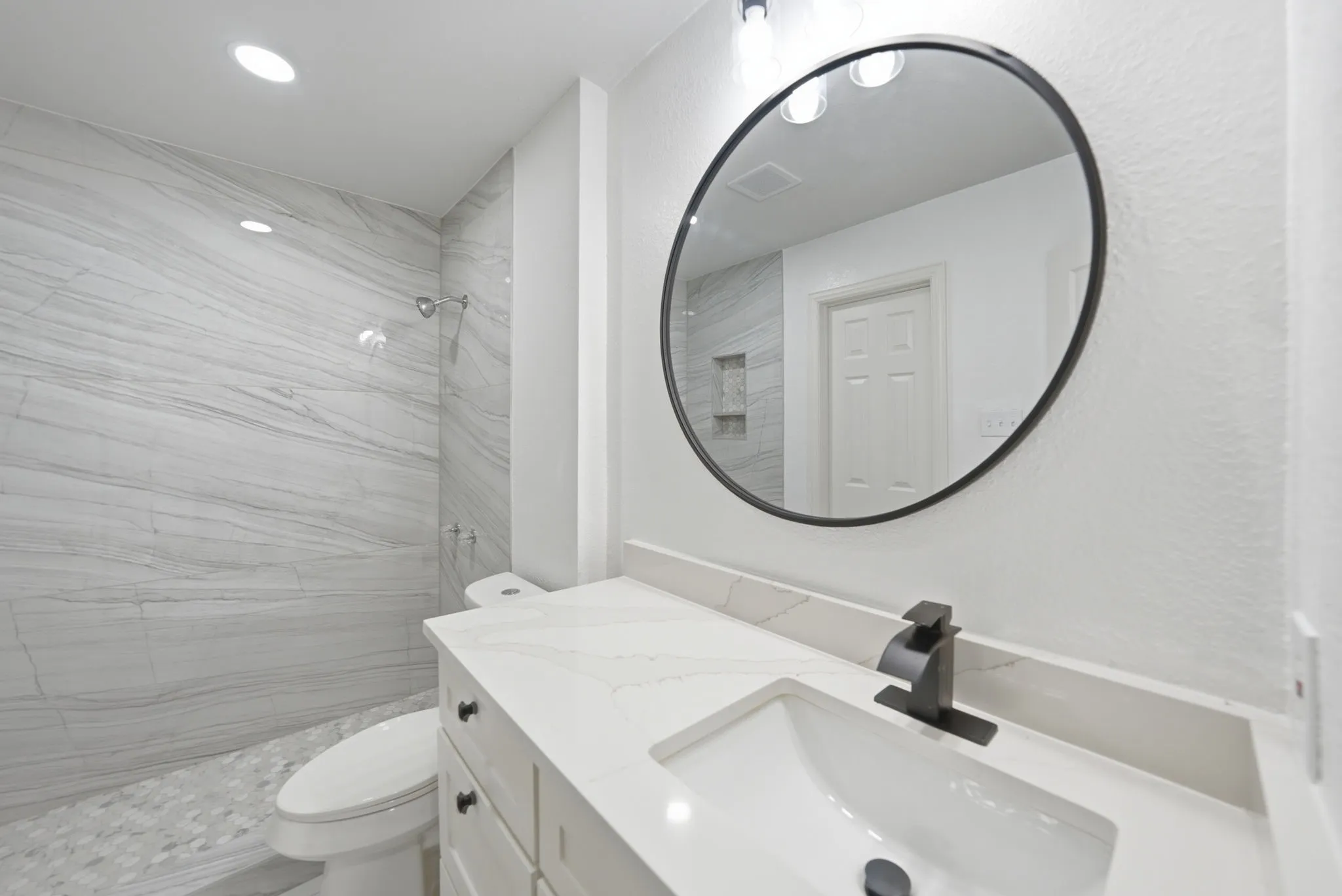 Walk-in shower with large format tile walls and pebble tile flooring, white vanity with stone countertop, round black-framed mirror, and matte black plumbing fixtures