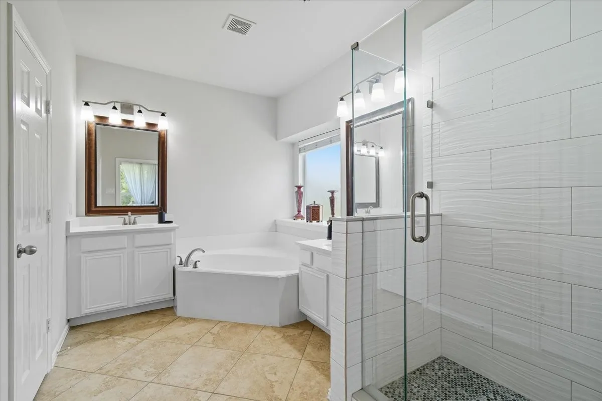 Dual vanity sinks with white cabinetry, corner soaking tub, glass-enclosed shower with mosaic tile floor, and large format wall tile