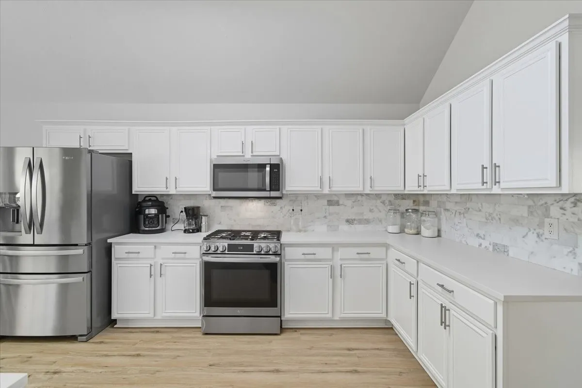 White cabinetry with brushed nickel hardware, stainless steel appliances, light-toned countertops, tile backsplash, and wood-finish flooring