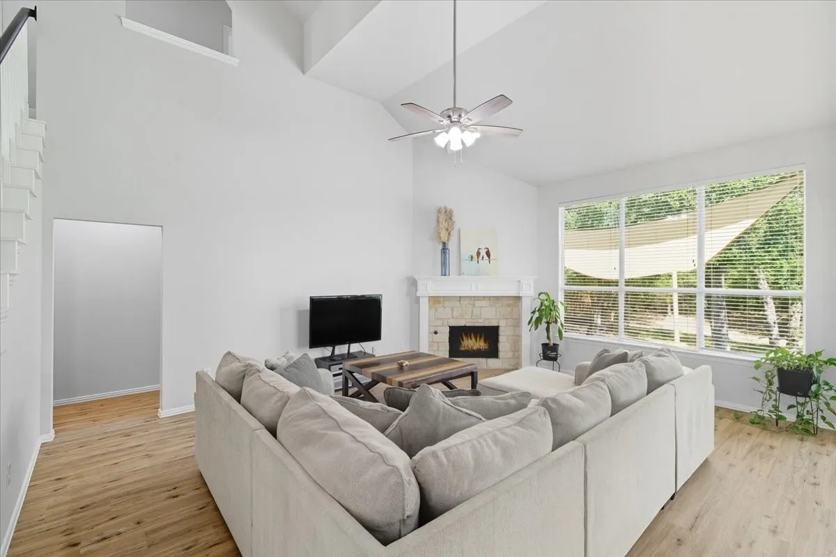 Vaulted ceiling, wood-finish flooring, stone-clad fireplace, large picture windows, and a ceiling fan with integrated lighting
