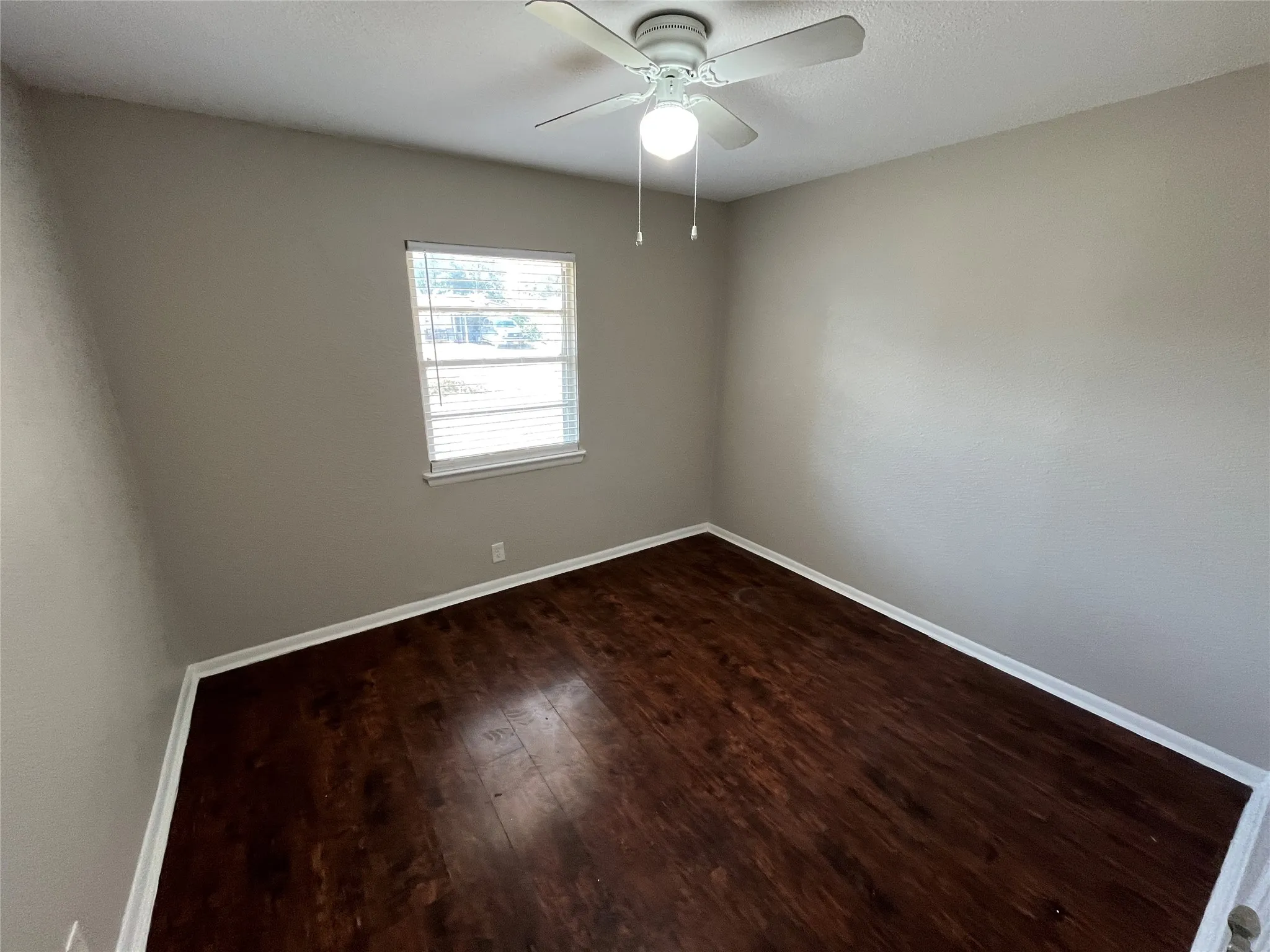 Bright, neutral bedroom featuring rich wood-finish flooring, soft natural light from a large window, and a ceiling fan with integrated lighting