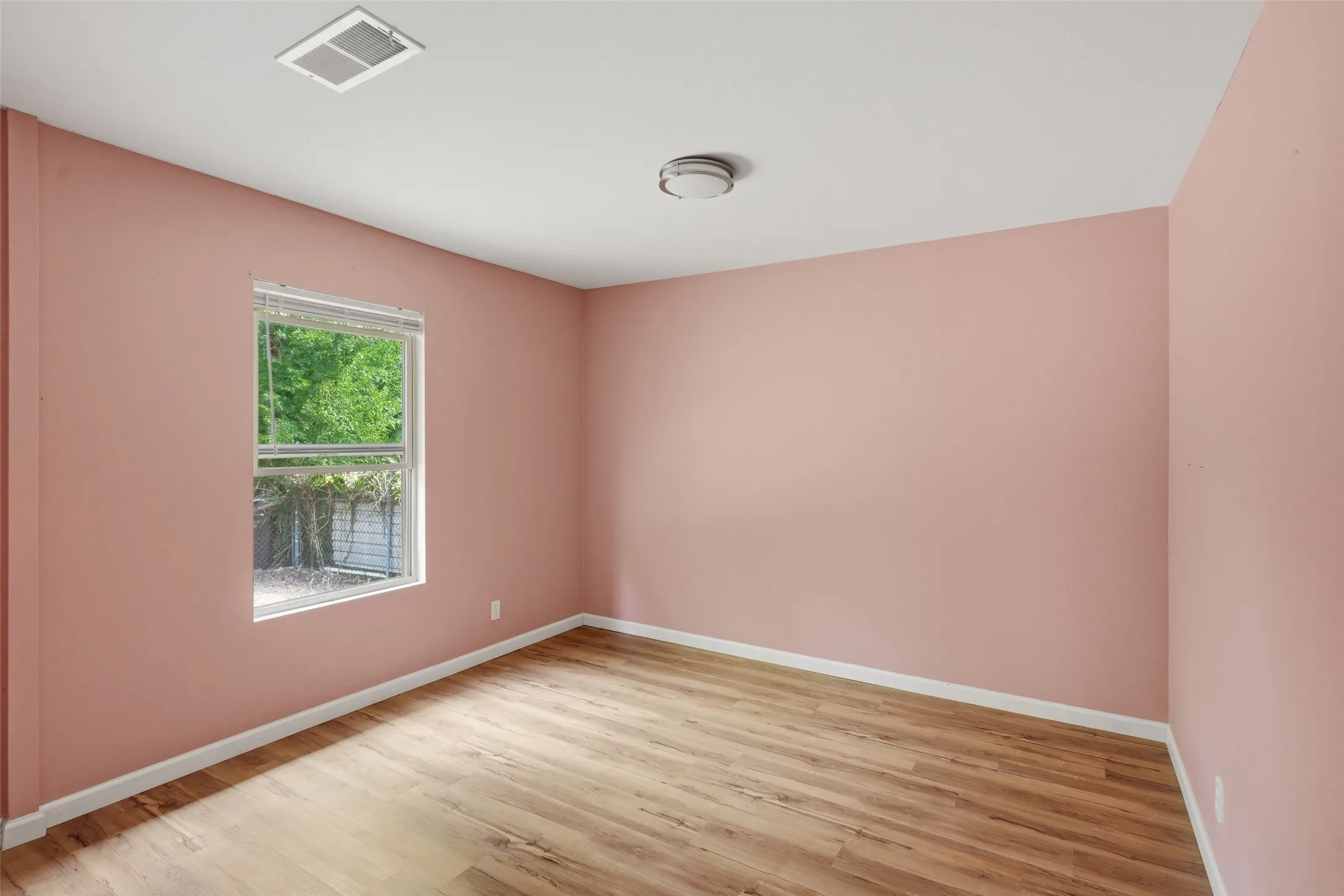 Bright and spacious bedroom featuring warm rose-toned walls, wood-finish flooring, and abundant natural light from a large double-hung window overlooking mature greenery