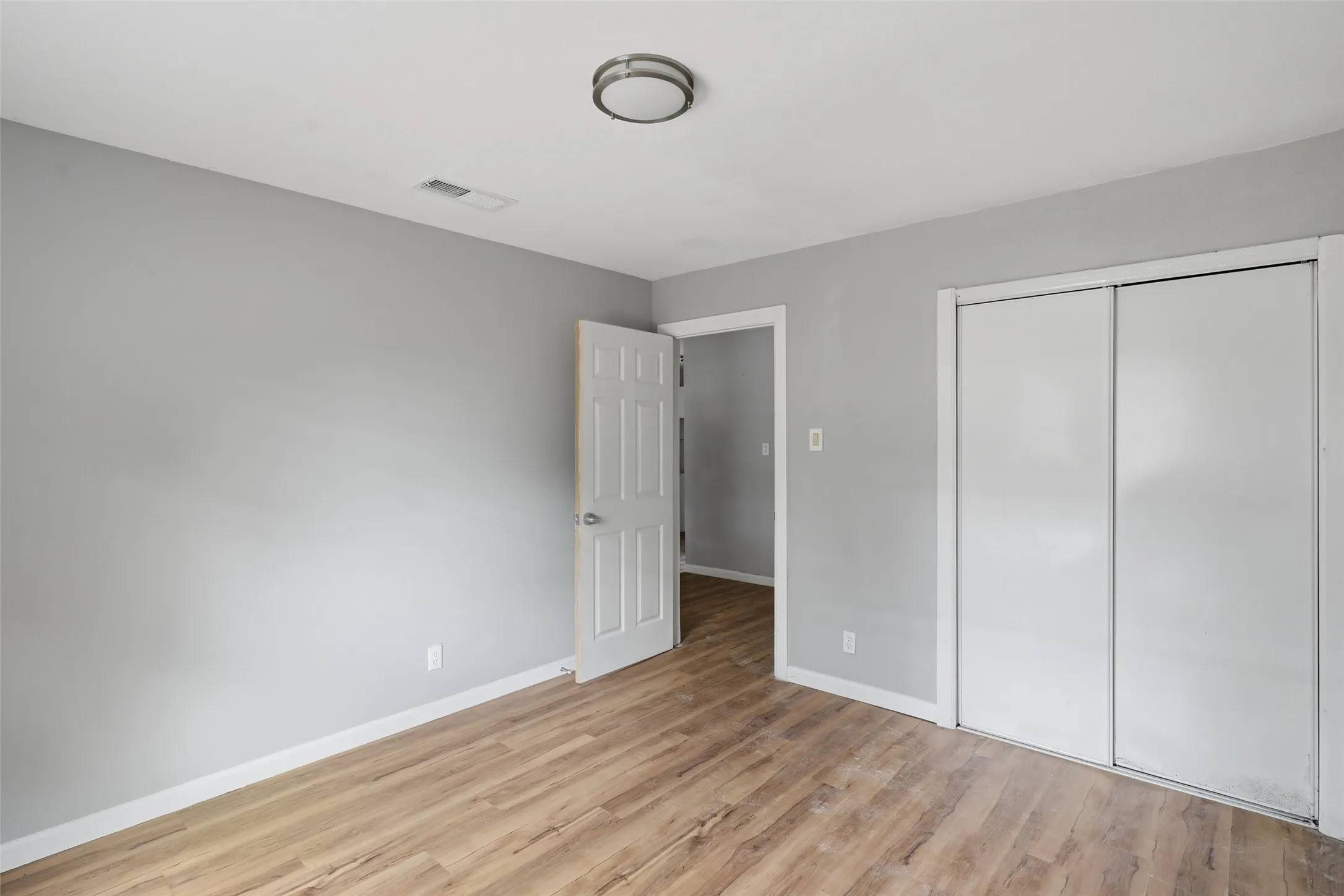 Wood-finish flooring, light gray wall paint, flush-mount ceiling light fixture, white interior door, and a two-panel sliding closet door