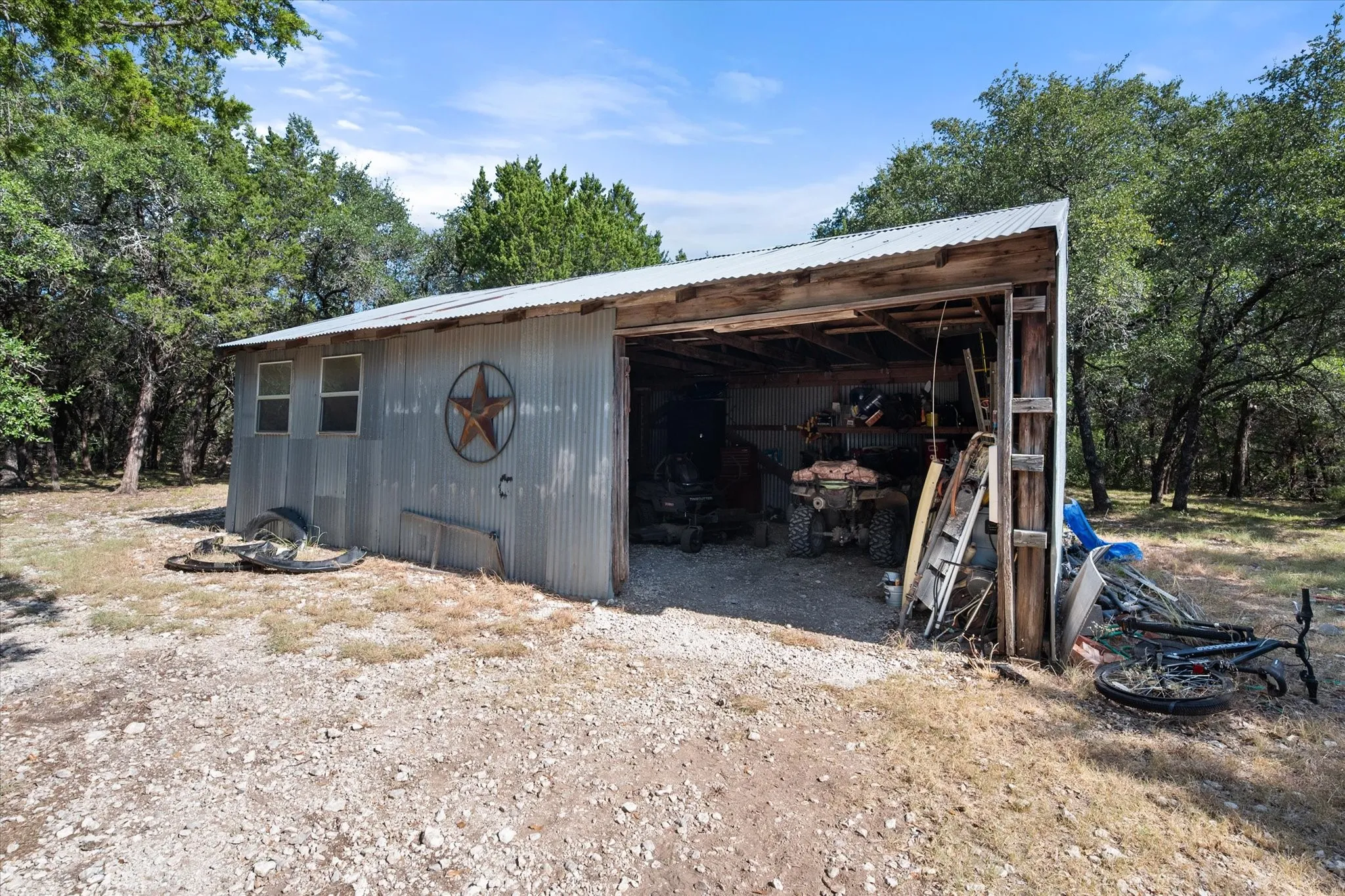 Corrugated metal outbuilding with a partially open front, featuring a peaked metal roof, exposed wood beam construction, and two small windows