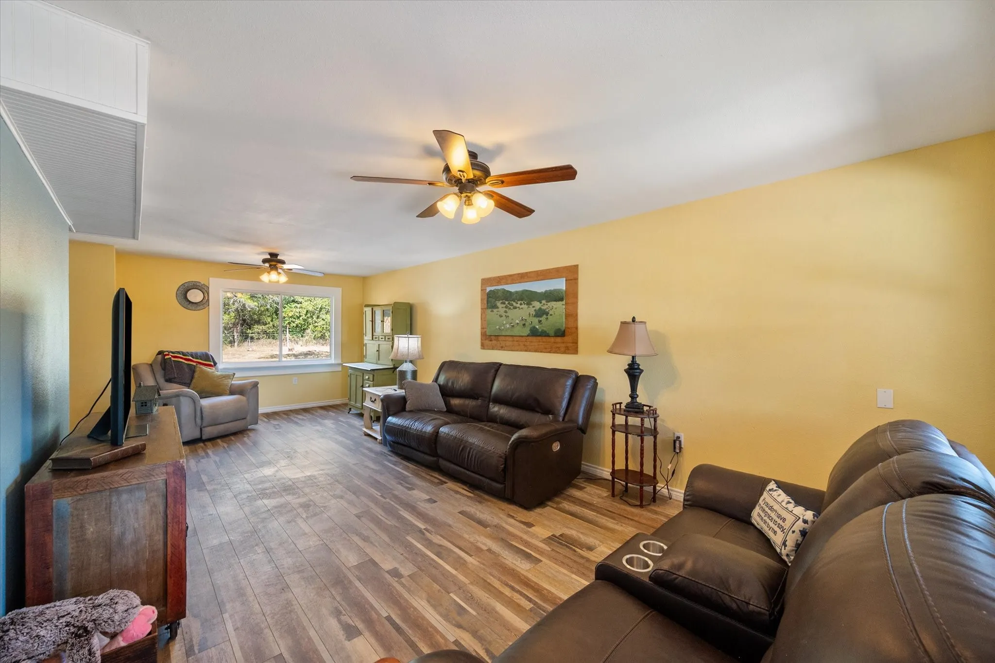 Spacious living area featuring wood-finish flooring, a window with natural light, and two ceiling fans with integrated lighting