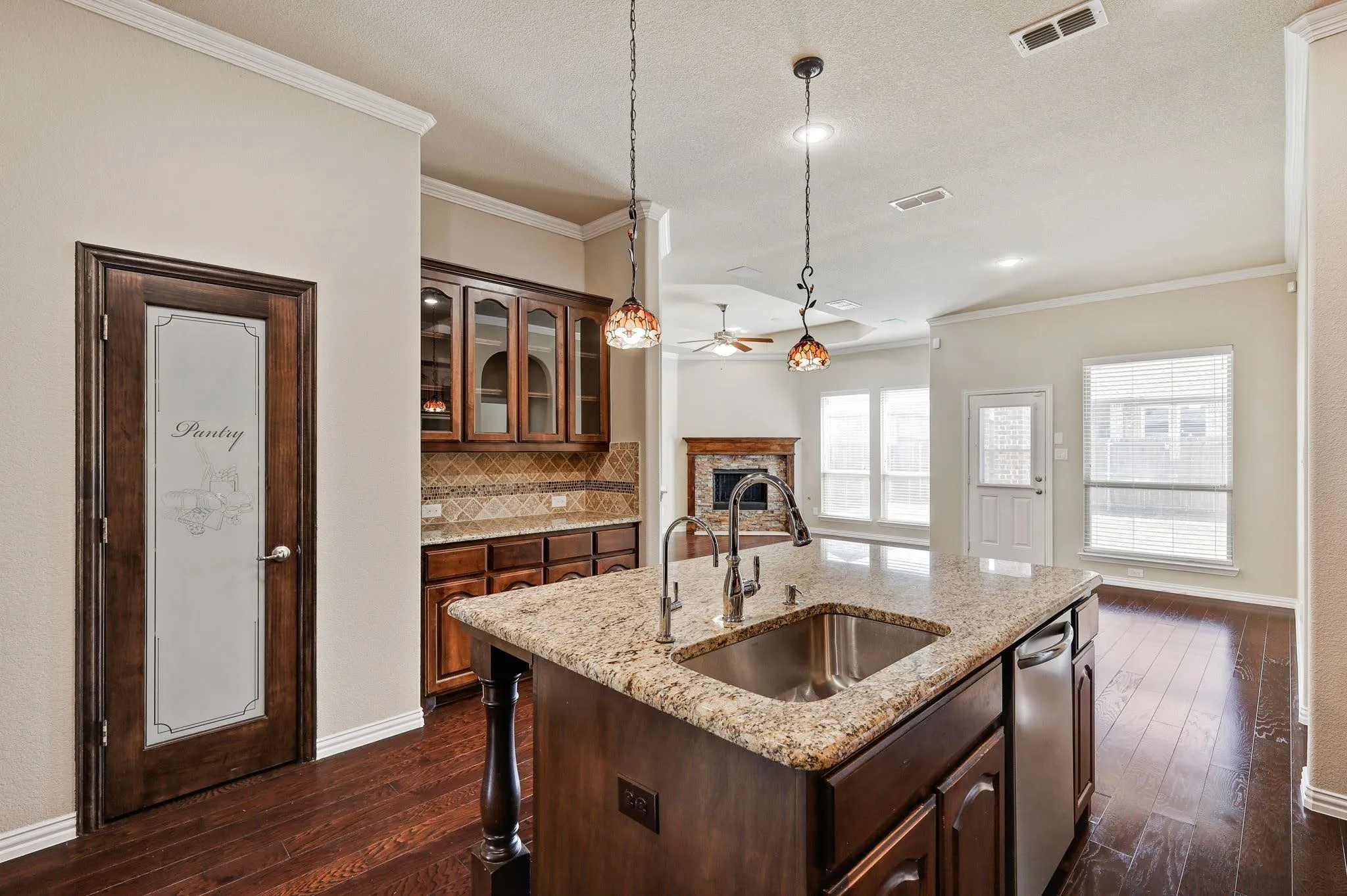 Kitchen island with a built-in sink and granite countertop, wood-finish flooring, dark wood cabinetry with glass-front uppers, and a pantry door with frosted glass