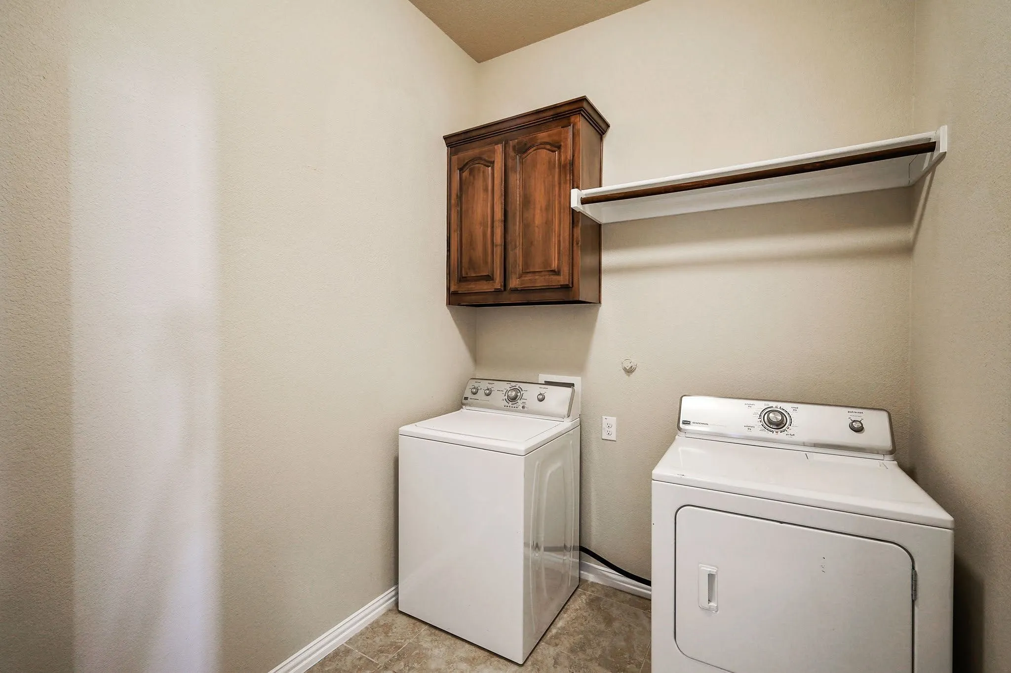 Laundry area featuring a wood-finish overhead cabinet, a white wall shelf, and tiled flooring