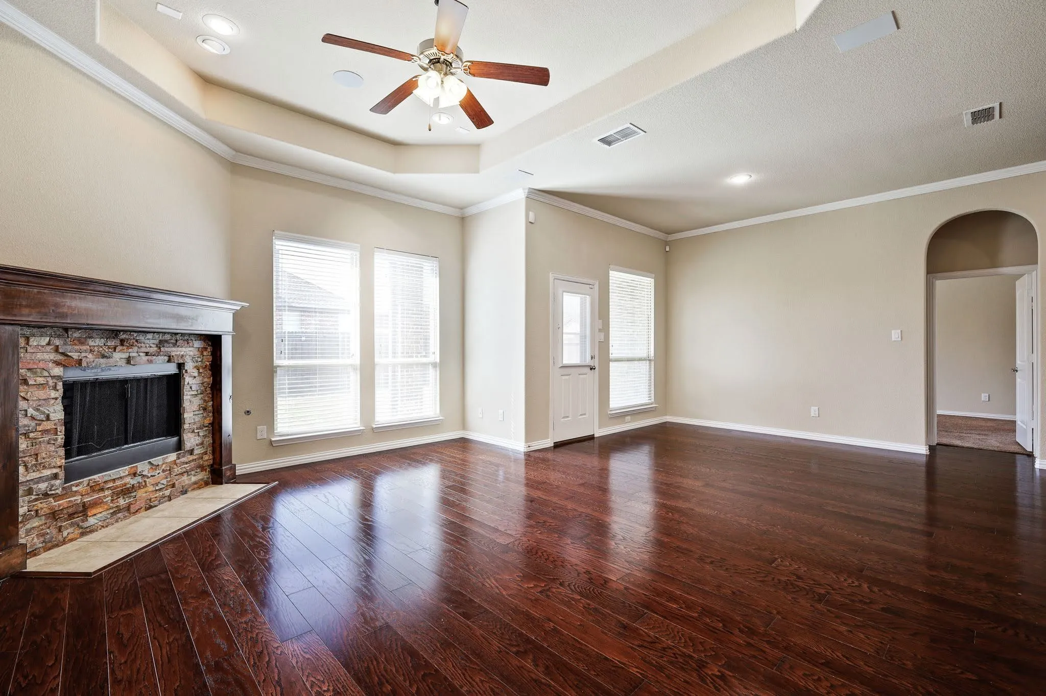 Hardwood-finish flooring, stone-clad fireplace with mantel, tray ceiling, recessed lighting, and ceiling fan