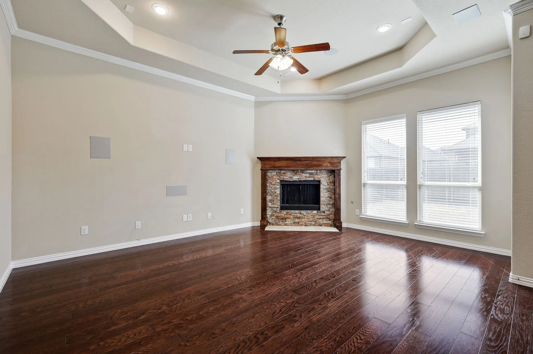 Tray ceiling with recessed lighting and a ceiling fan, wood-finish flooring, stone-clad fireplace with a wood mantel, and multiple windows