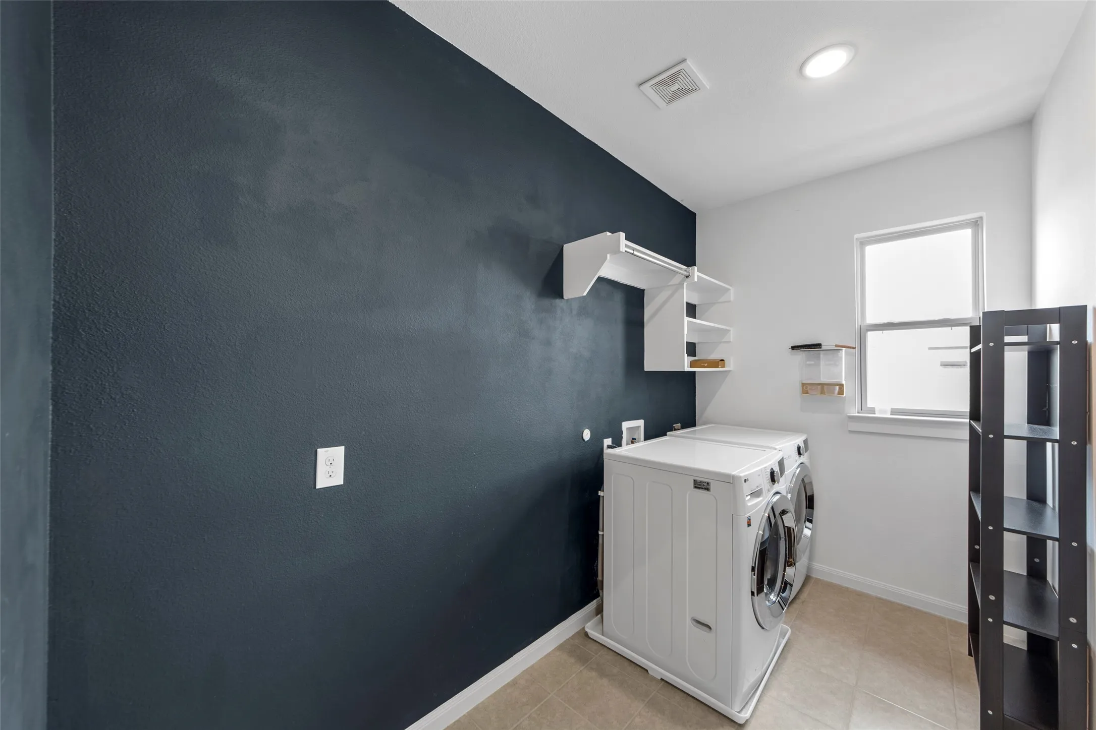 Modern laundry room featuring a front-load washer and dryer combination, white cabinetry with integrated storage, a utility sink with chrome faucet, dark accent wall, and abundant natural light from a large window