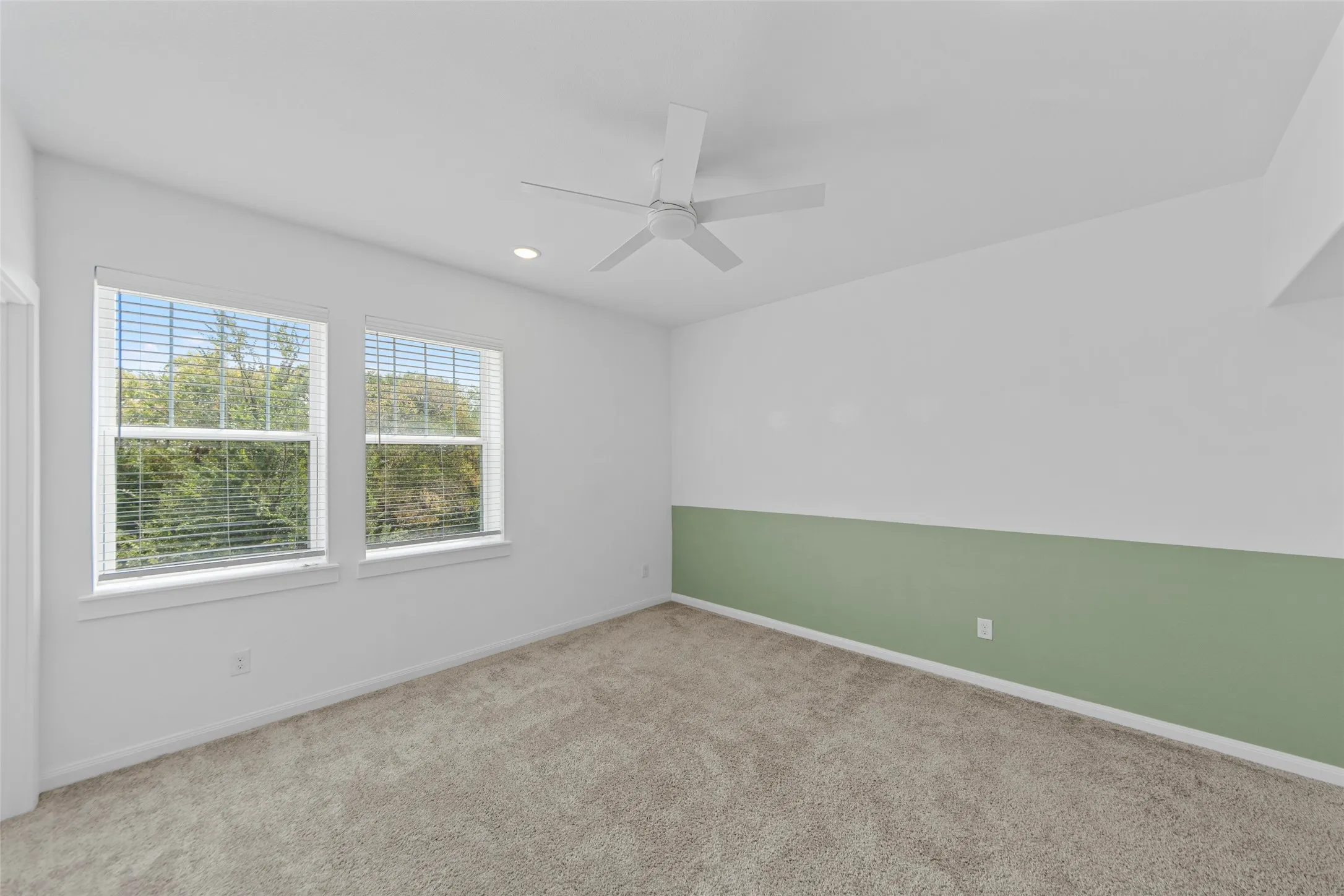 Bright secondary bedroom showcasing abundant natural light from dual casement windows, neutral carpet flooring, ceiling fan with integrated lighting, and a fresh two-tone paint treatment featuring soft sage green wainscoting against crisp white walls