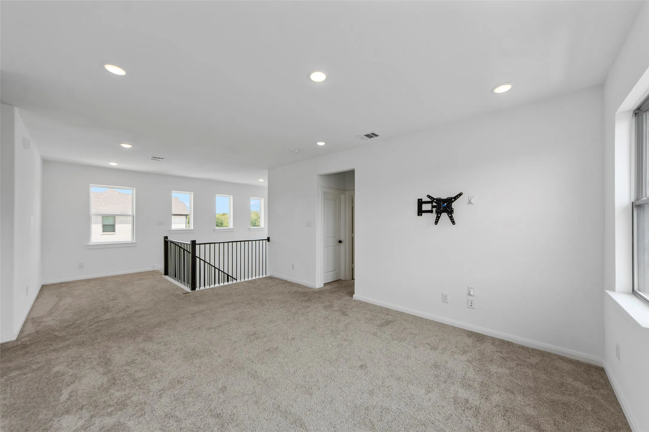 Neutral-toned carpeting, recessed lighting, multiple windows, white walls, and a black metal spindle railing
