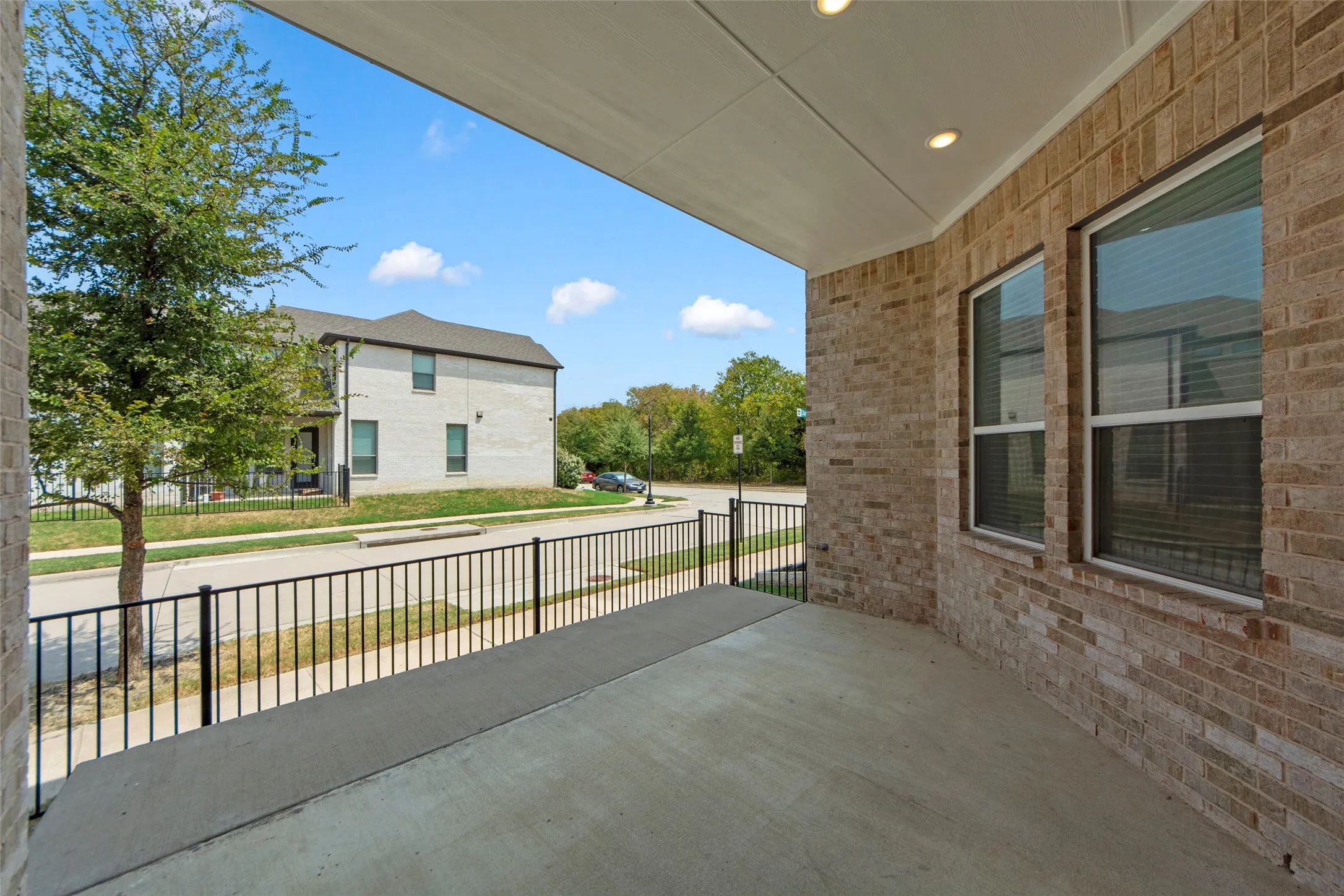 Covered concrete patio featuring black metal railing with horizontal spindles, brick exterior wall, and large multi-pane windows