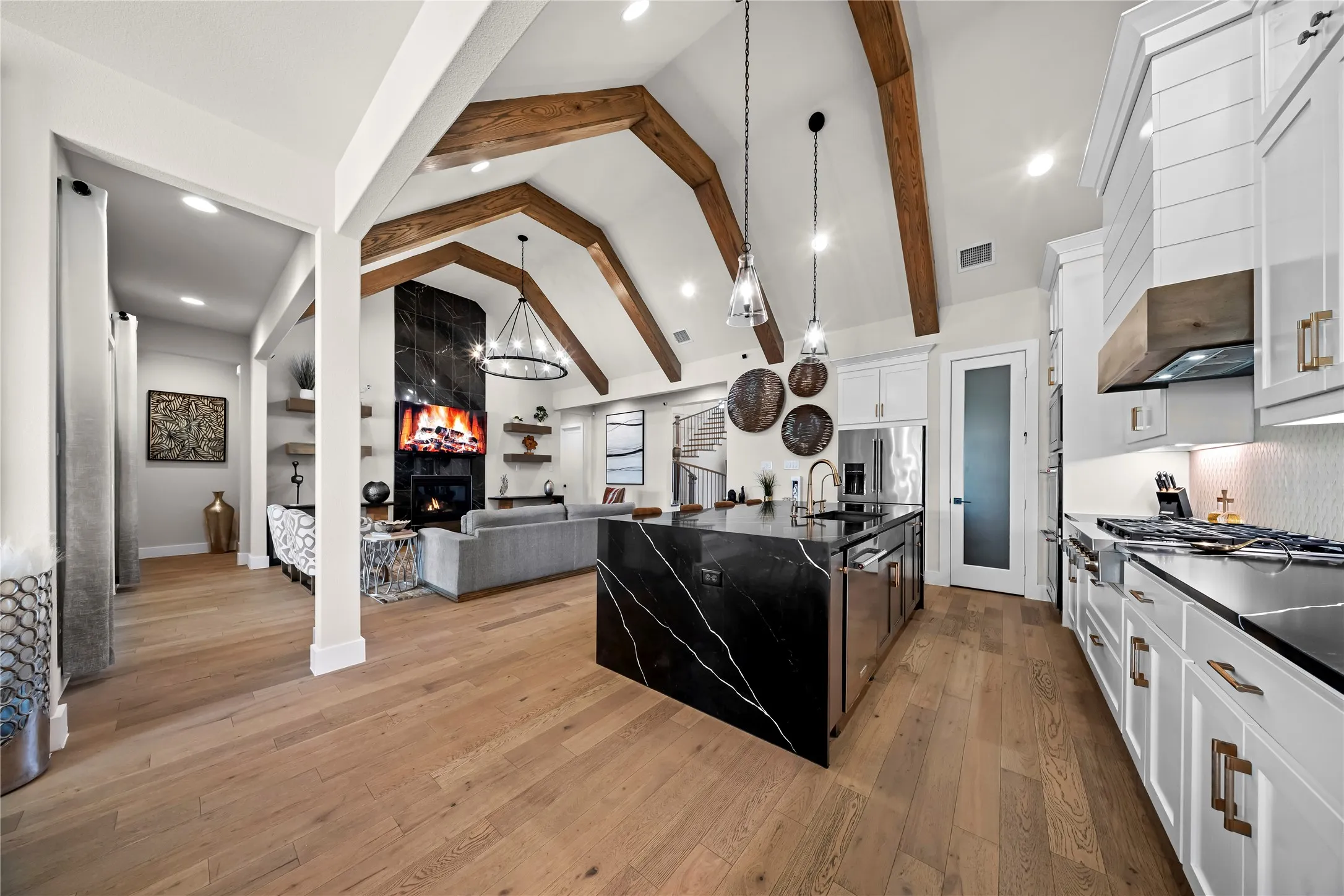 Vaulted ceilings with exposed wood beams, wood-finish flooring, black stone kitchen island with waterfall edge, white shaker cabinetry, and a black stone fireplace surround