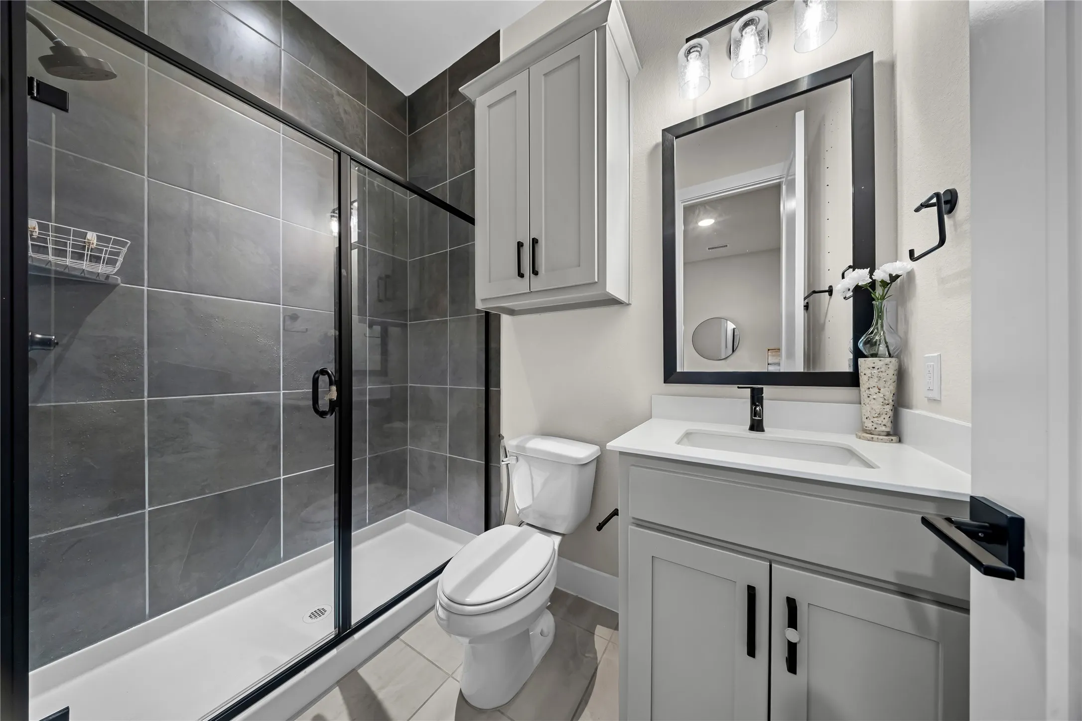 Walk-in shower with dark tile surround, light gray vanity with white countertop, and framed mirror with integrated lighting