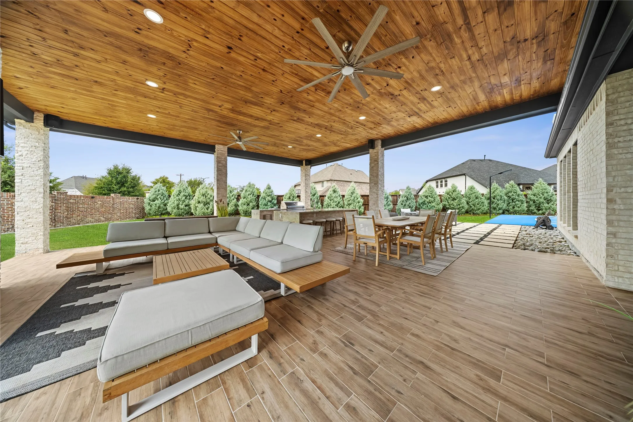 Expansive covered patio featuring a wood-panel ceiling with recessed lighting and ceiling fans, stone-clad support columns, wood-finish tile flooring, and a built-in outdoor kitchen area